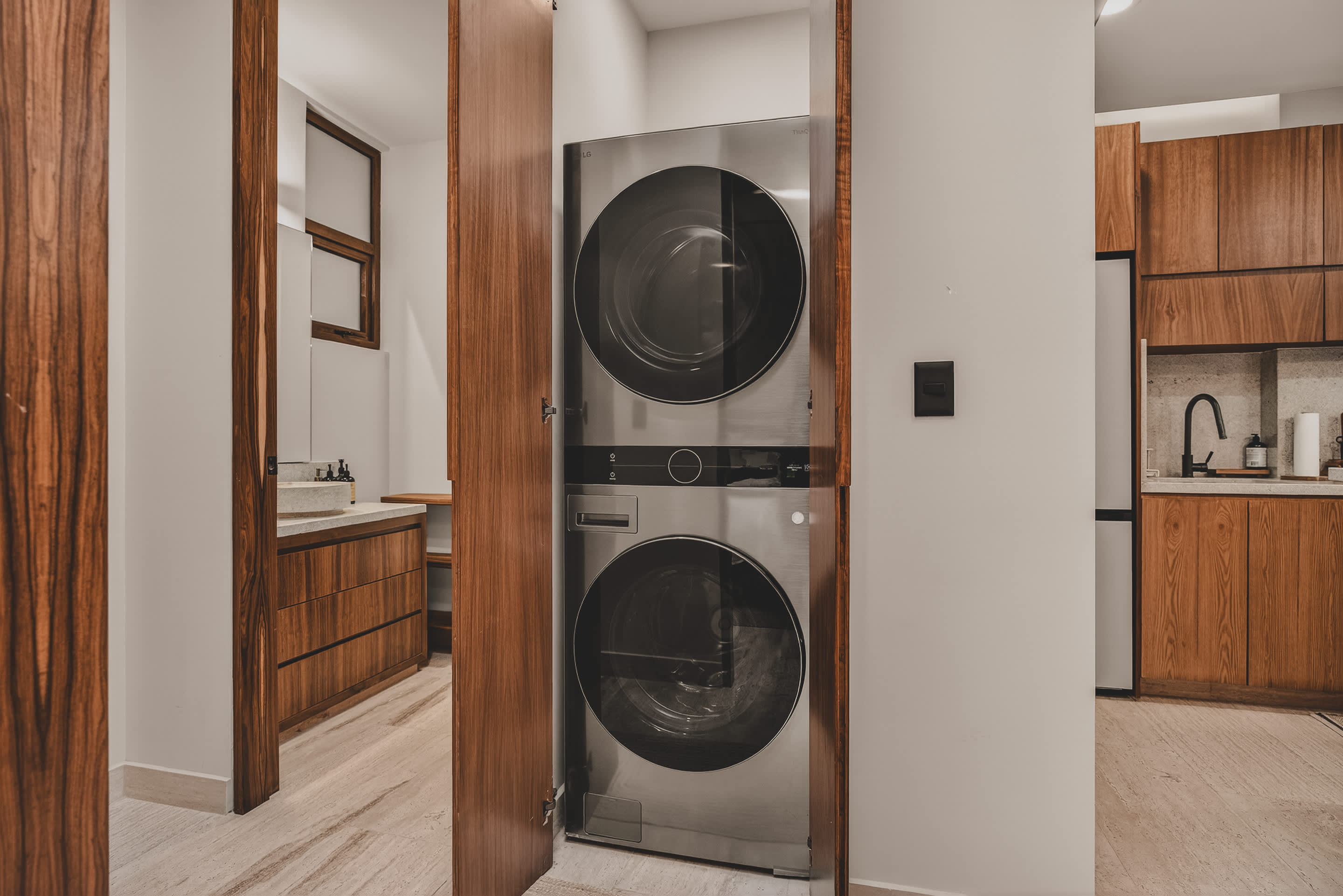 A stacked silver washer and dryer unit is neatly tucked away in a closet with wooden bifold doors, adjacent to a modern bathroom vanity visible through an open doorway.