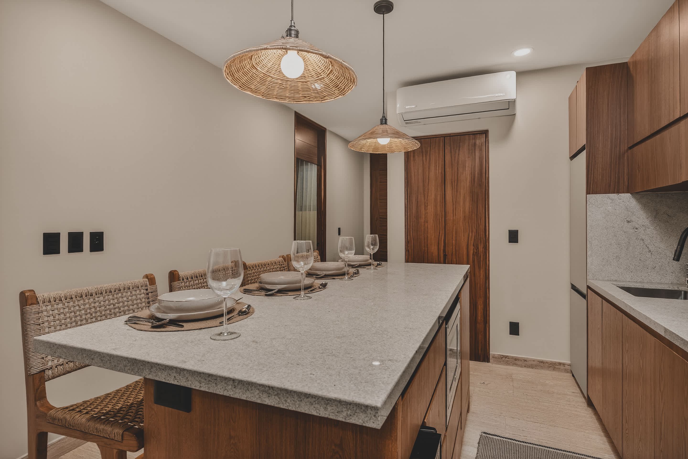 A modern dining area features a light speckled granite island with four place settings, illuminated by two woven pendant lights, and backed by light beige walls and wooden cabinetry.