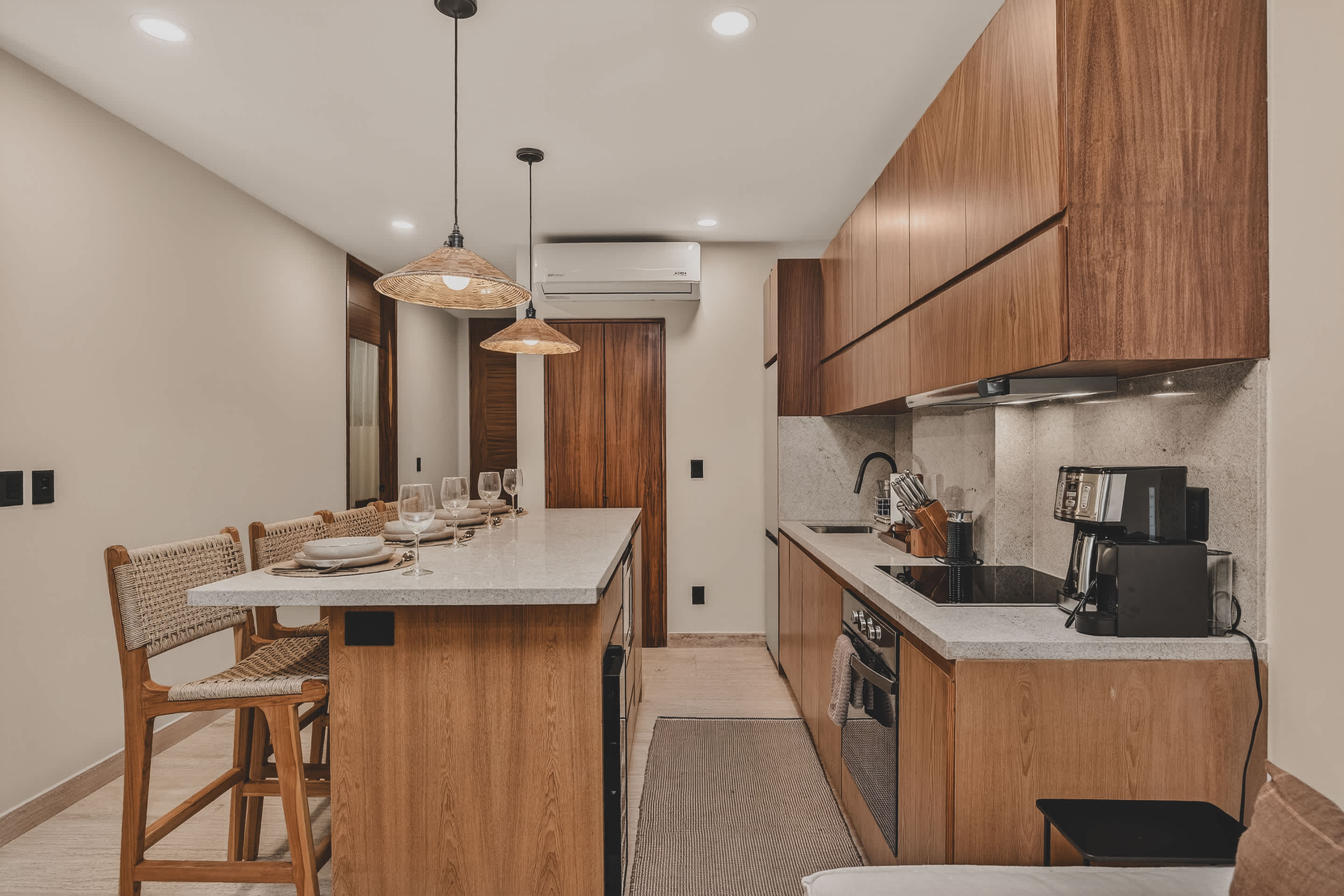 A contemporary kitchen and dining space showcases a light speckled island with four woven bar stools, prepared for a meal, illuminated by two woven pendant lights. The kitchen wall features extensive dark wood cabinetry and a built-in refrigerator.