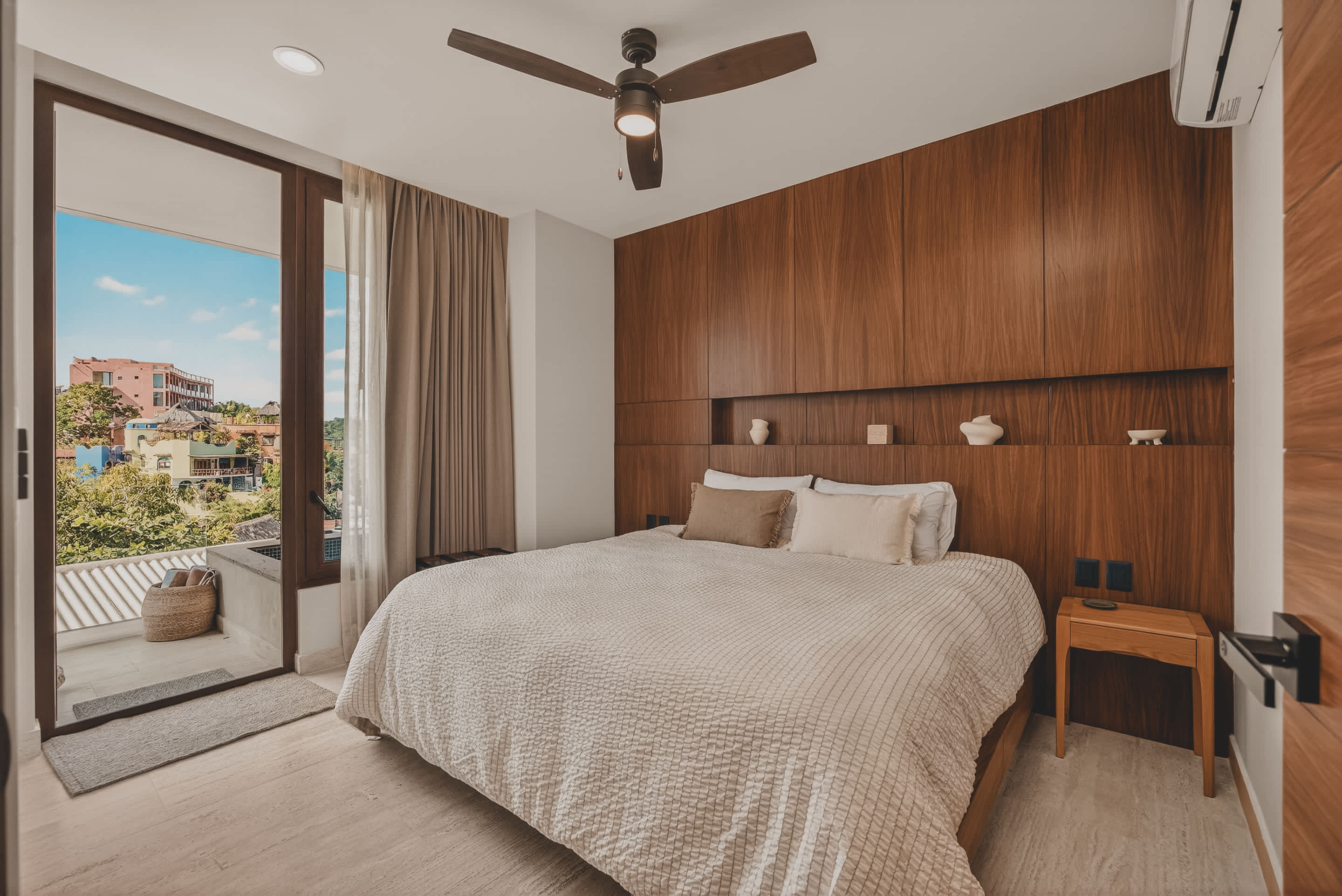 A modern bedroom features a king-sized bed with light bedding and a dark wooden headboard integrated into a full accent wall with shelving. Large glass doors open to a private balcony with a view, and a ceiling fan hangs overhead.