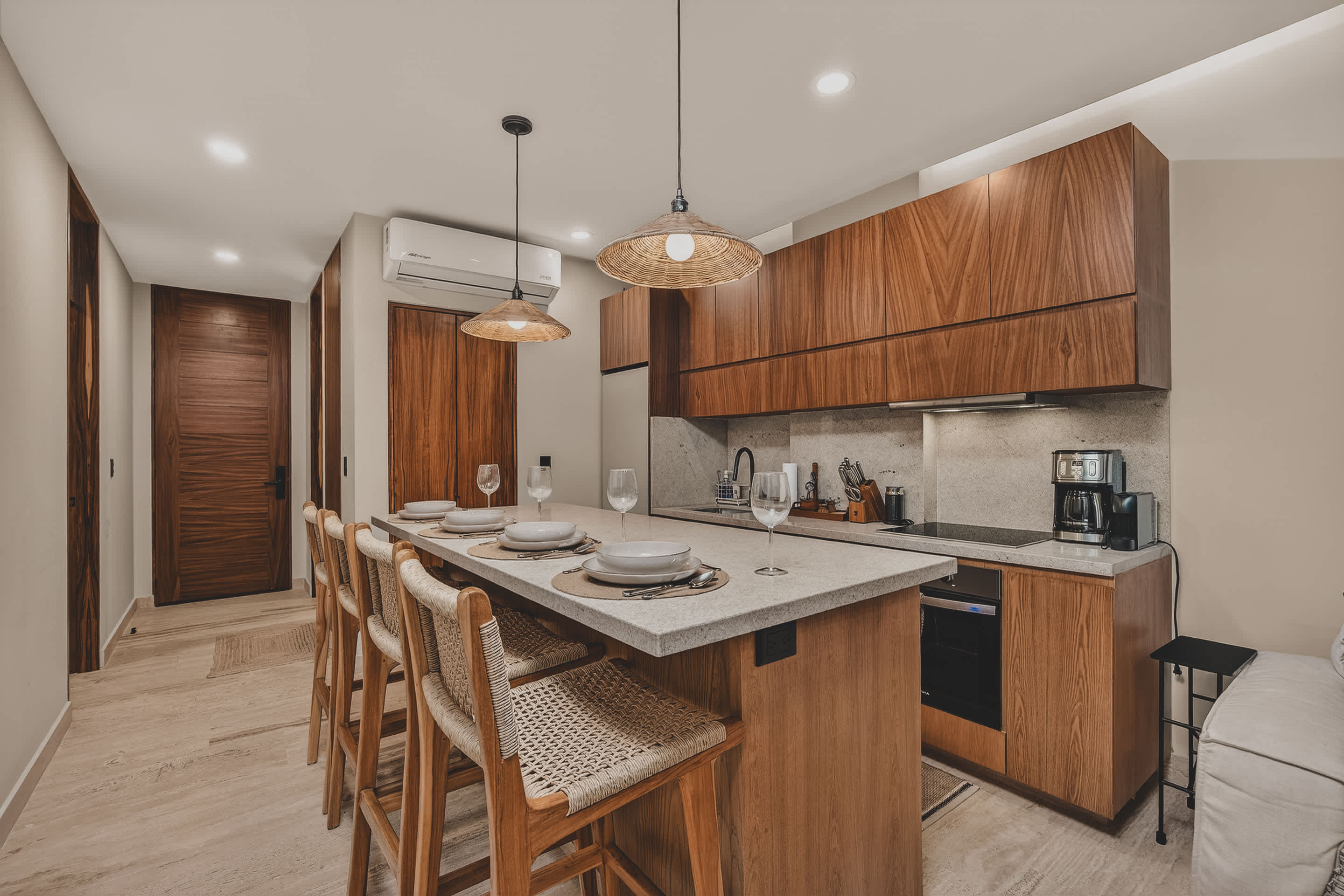 A modern kitchen and dining area features a light speckled island with four woven bar stools, set with dinnerware, under two woven pendant lights. Dark wood cabinetry lines the wall, and a refrigerator is visible at the end of the counter.