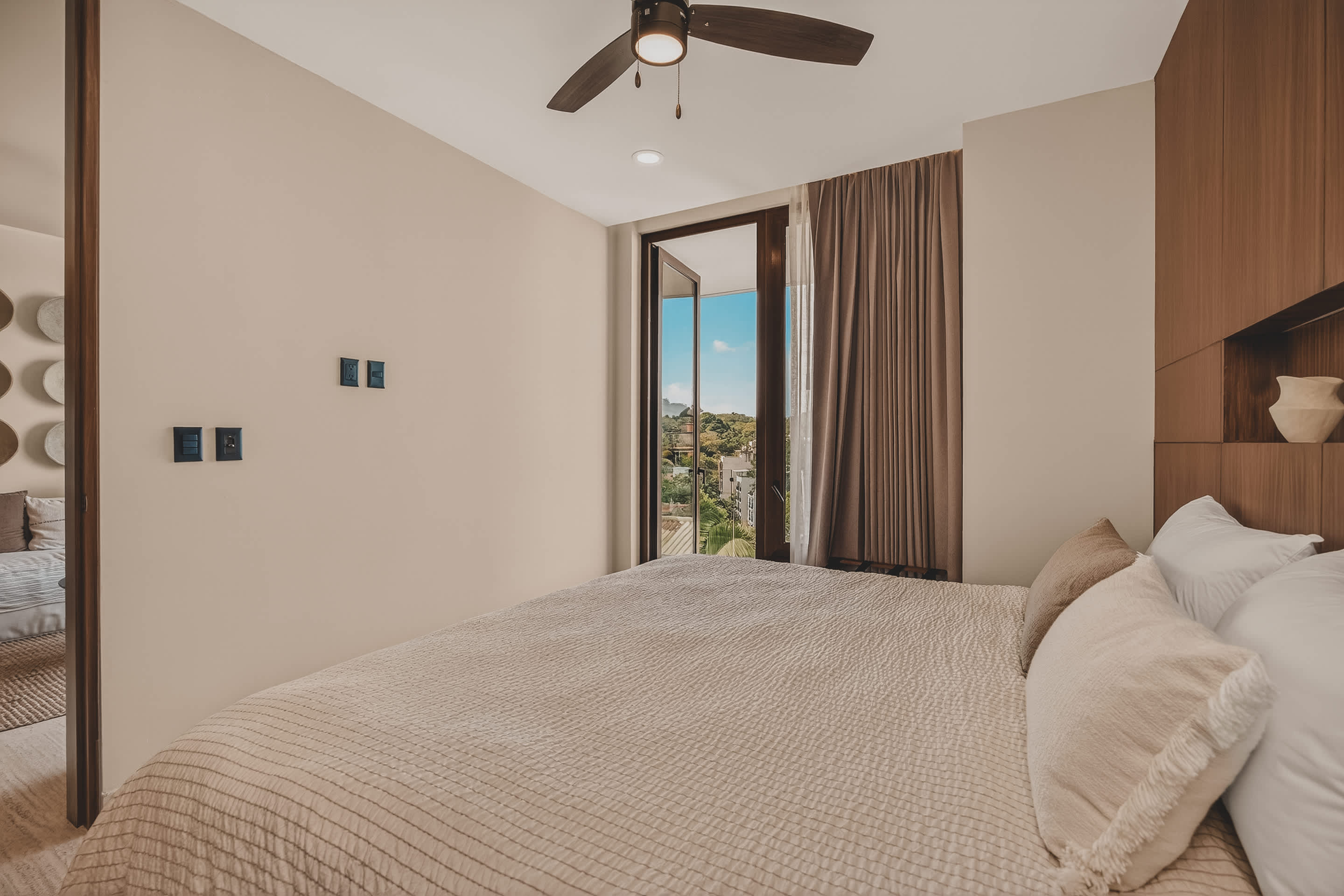 A bright bedroom features a king-sized bed with light-colored bedding and pillows, with a view of the outdoors through large glass doors leading to a balcony. The room has light-colored walls and a ceiling fan.