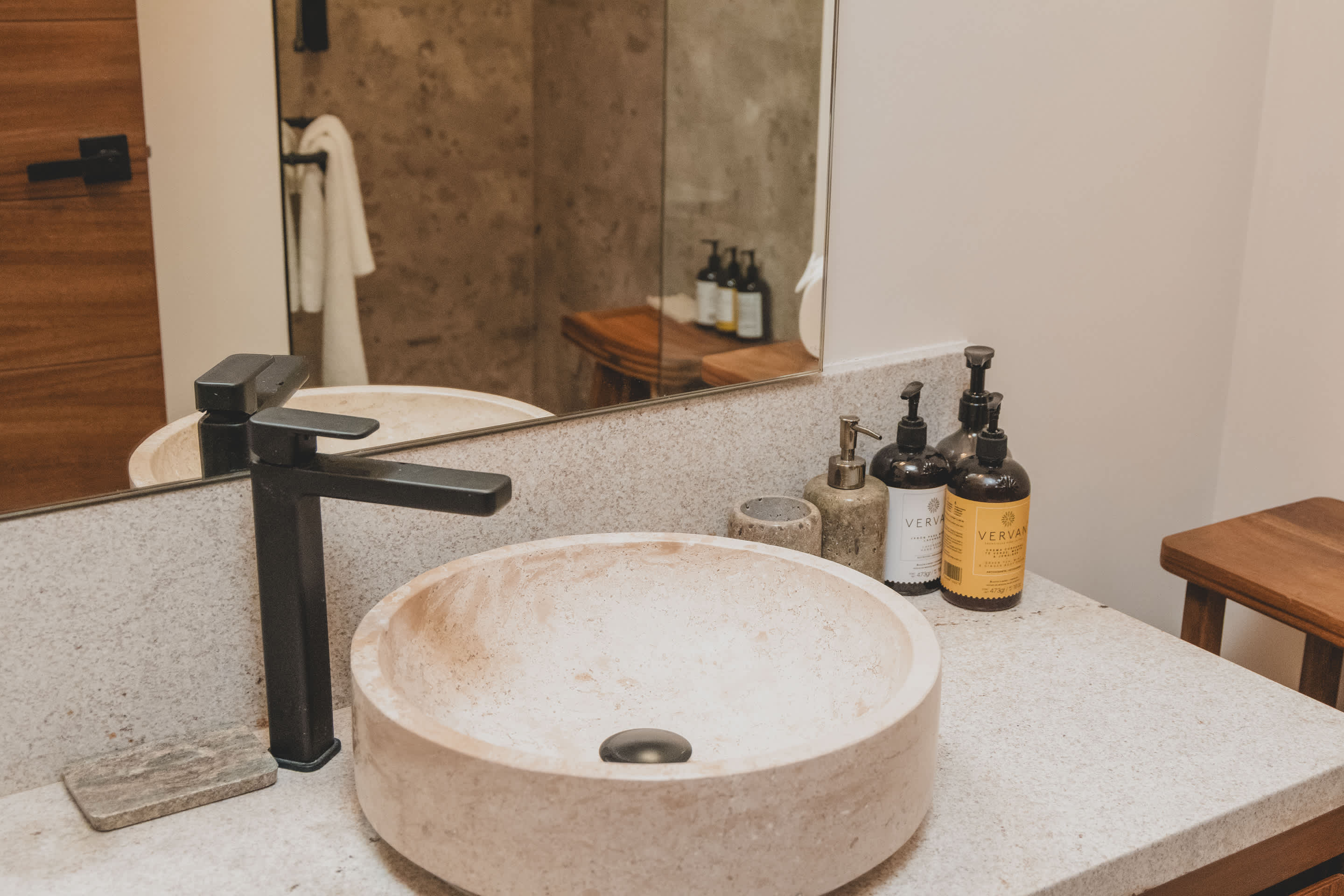 A modern bathroom vanity features a round, light-colored stone vessel sink with a matte black faucet, set on a speckled granite countertop, with a large mirror reflecting wooden elements.