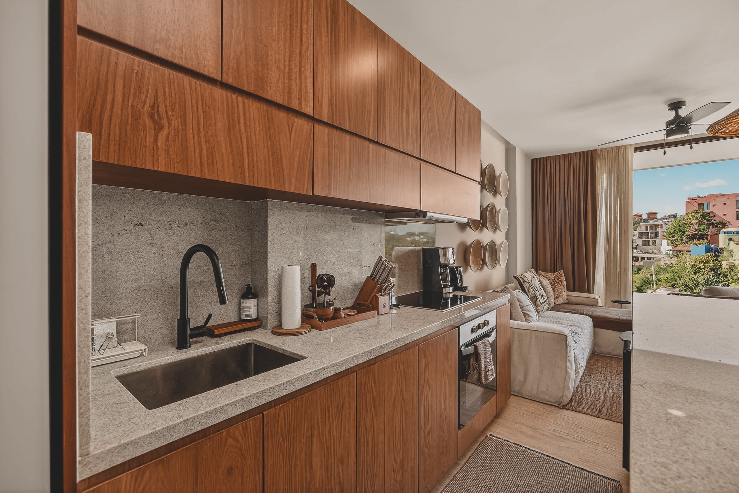 A sleek kitchen counter features a black sink and faucet, light speckled backsplash, and dark wood upper and lower cabinetry. A coffee maker, knife block, and various kitchen essentials are neatly arranged on the counter.
