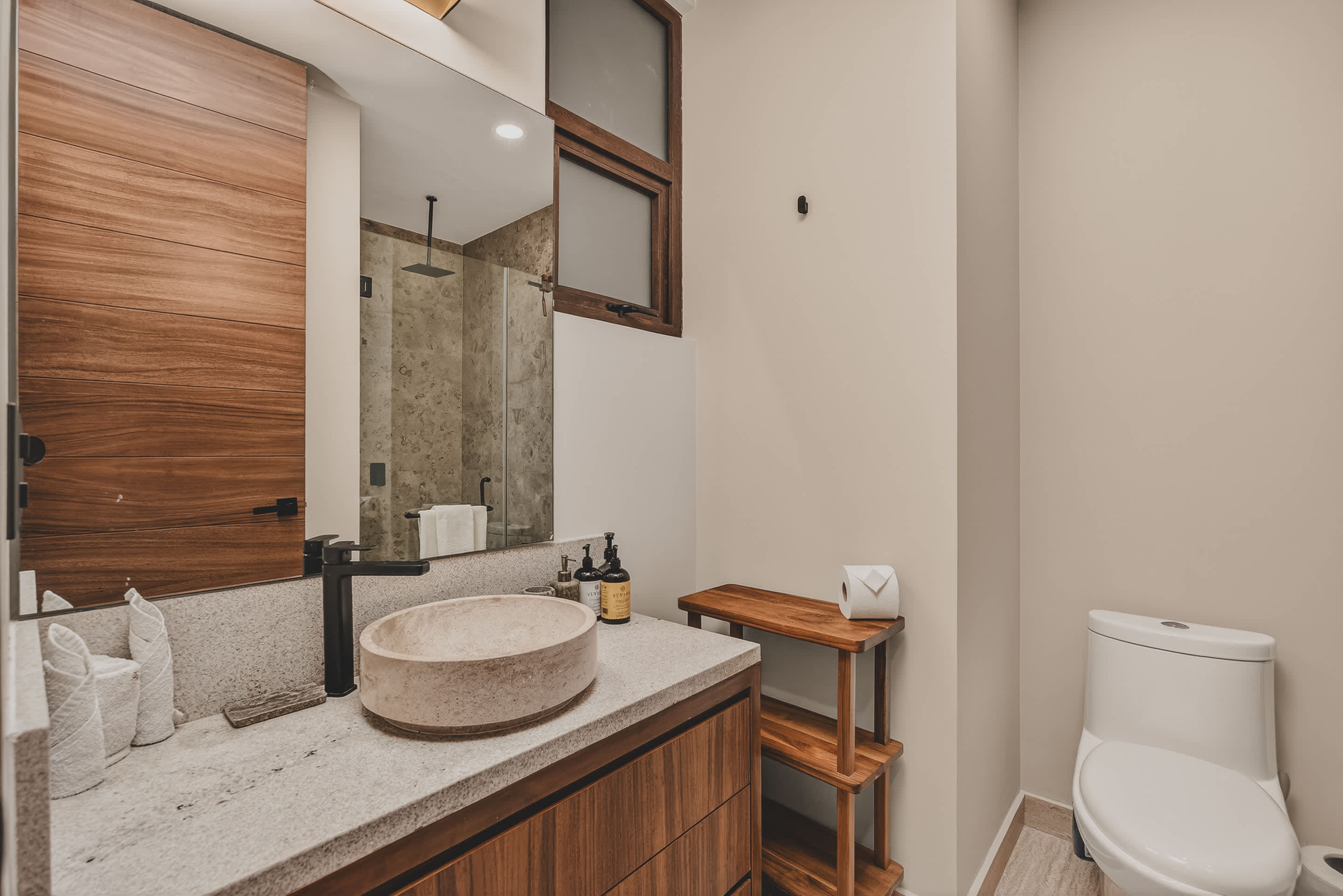 Modern bathroom featuring a floating wooden vanity with a light-colored stone countertop and a round vessel sink, a large mirror, and a toilet, with a wooden stool holding toilet paper.