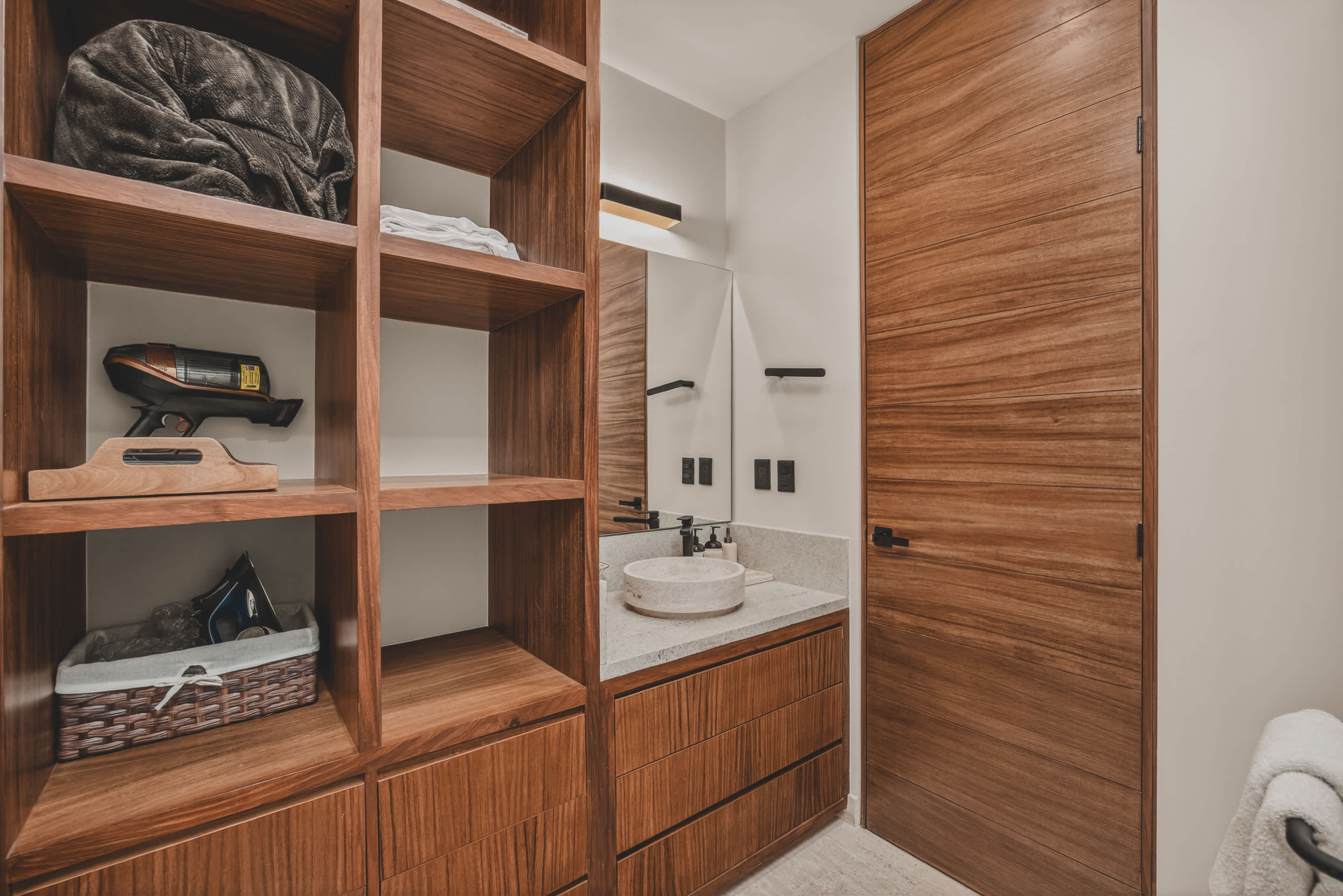 A modern bathroom vanity area features a light-colored vessel sink on a speckled countertop, dark wood cabinetry, and open shelving filled with towels and a hairdryer. A full-height wooden door stands to the right, complementing the warm wood tones.