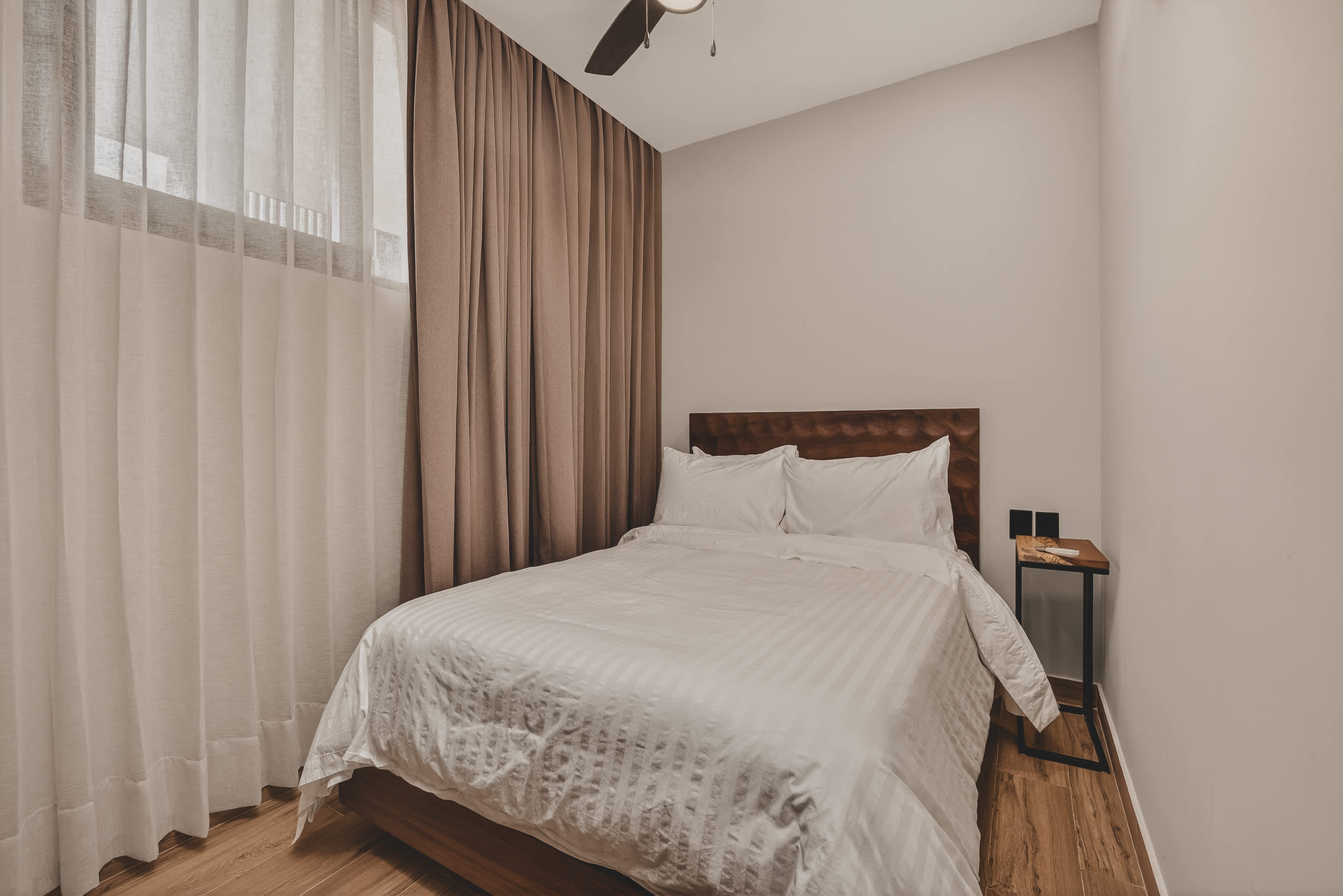 A comfortable bedroom featuring a queen bed with a dark wooden headboard, flanked by a small wooden bedside table. The room has light-colored curtains, a ceiling fan, and warm wooden flooring.