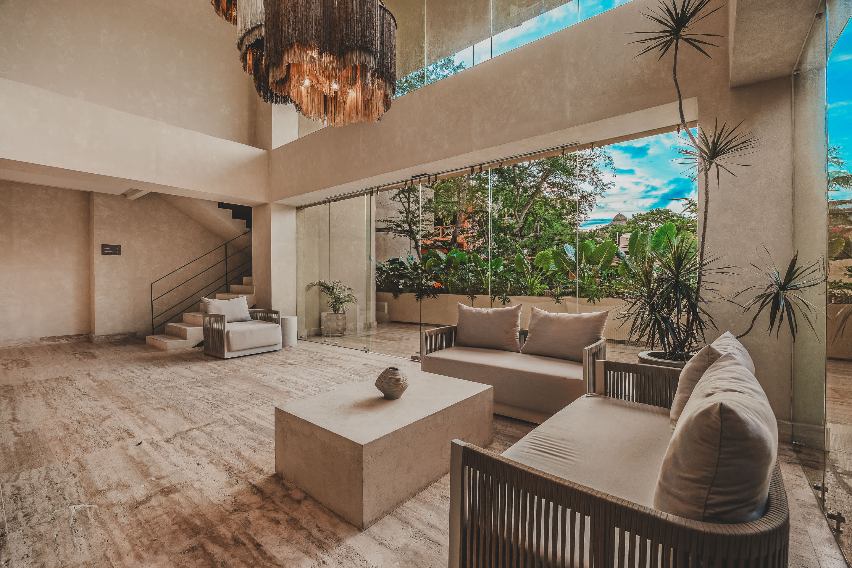A bright and airy lobby seating area with two modern sofas and an armchair, a concrete coffee table, and large glass walls overlooking lush tropical greenery. Decorative chandeliers hang from the high ceiling.