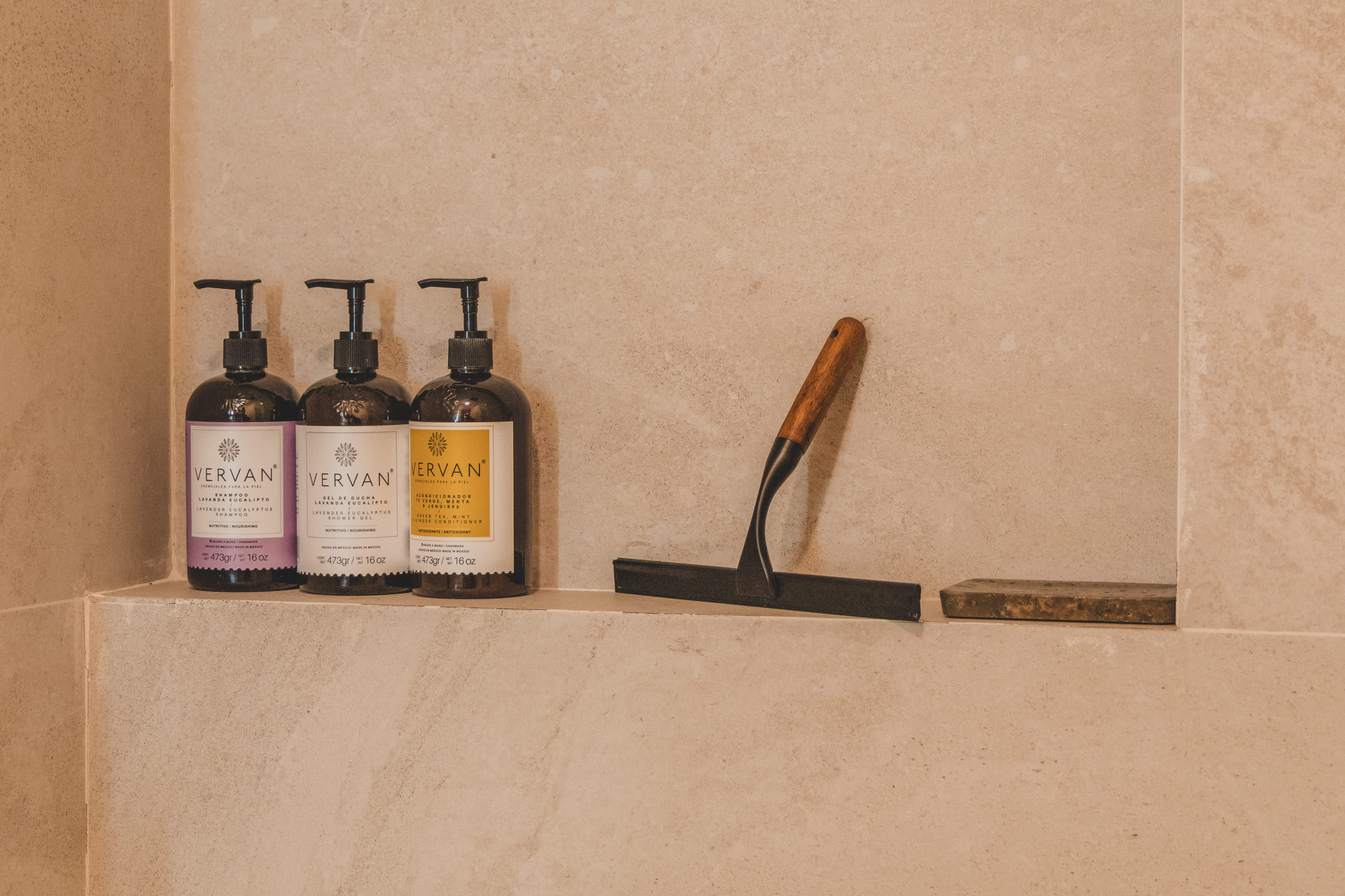 A tiled shower niche holds three pump bottles of 'VERVAN' brand toiletries in purple and yellow, alongside a shower squeegee with a wooden handle. The wall is covered in light-colored rectangular tiles.