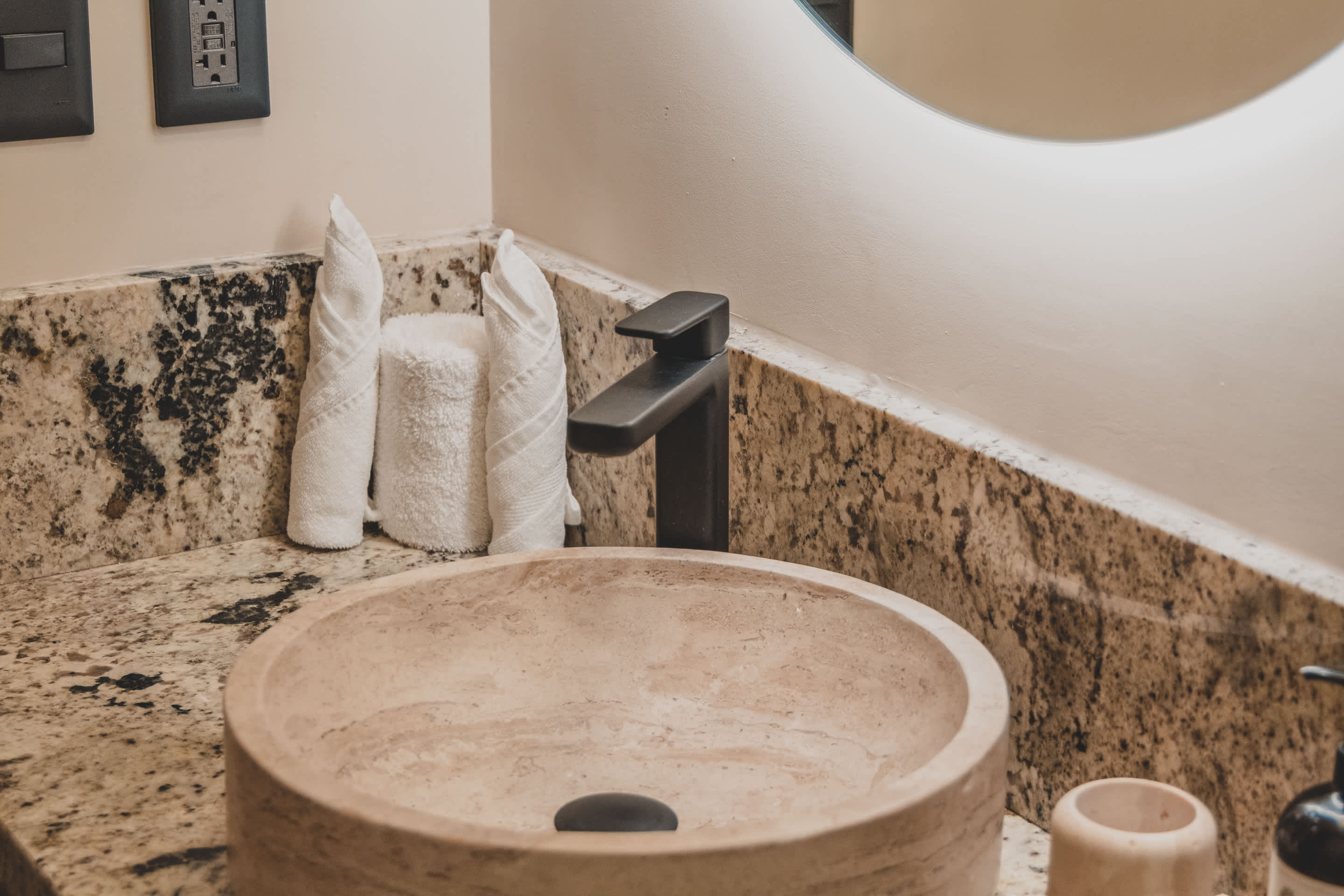 A close-up of a modern bathroom vanity featuring a round stone vessel sink and a matte black faucet, set on a granite countertop. Two white towels are decoratively folded, with a circular backlit mirror partially visible above.
