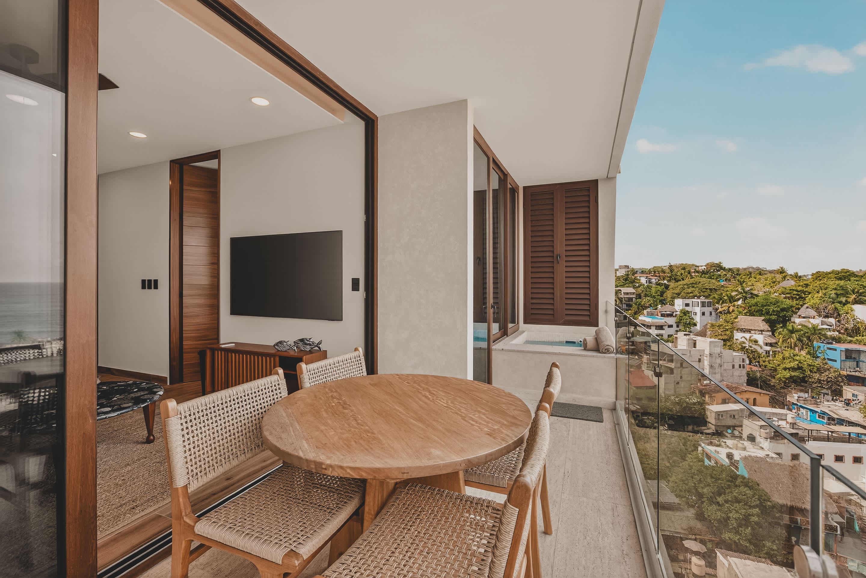 A wide-angle view from indoors showcases a private balcony with a wooden dining set and a built-in hot tub, all overlooking a picturesque coastal town and the ocean.
