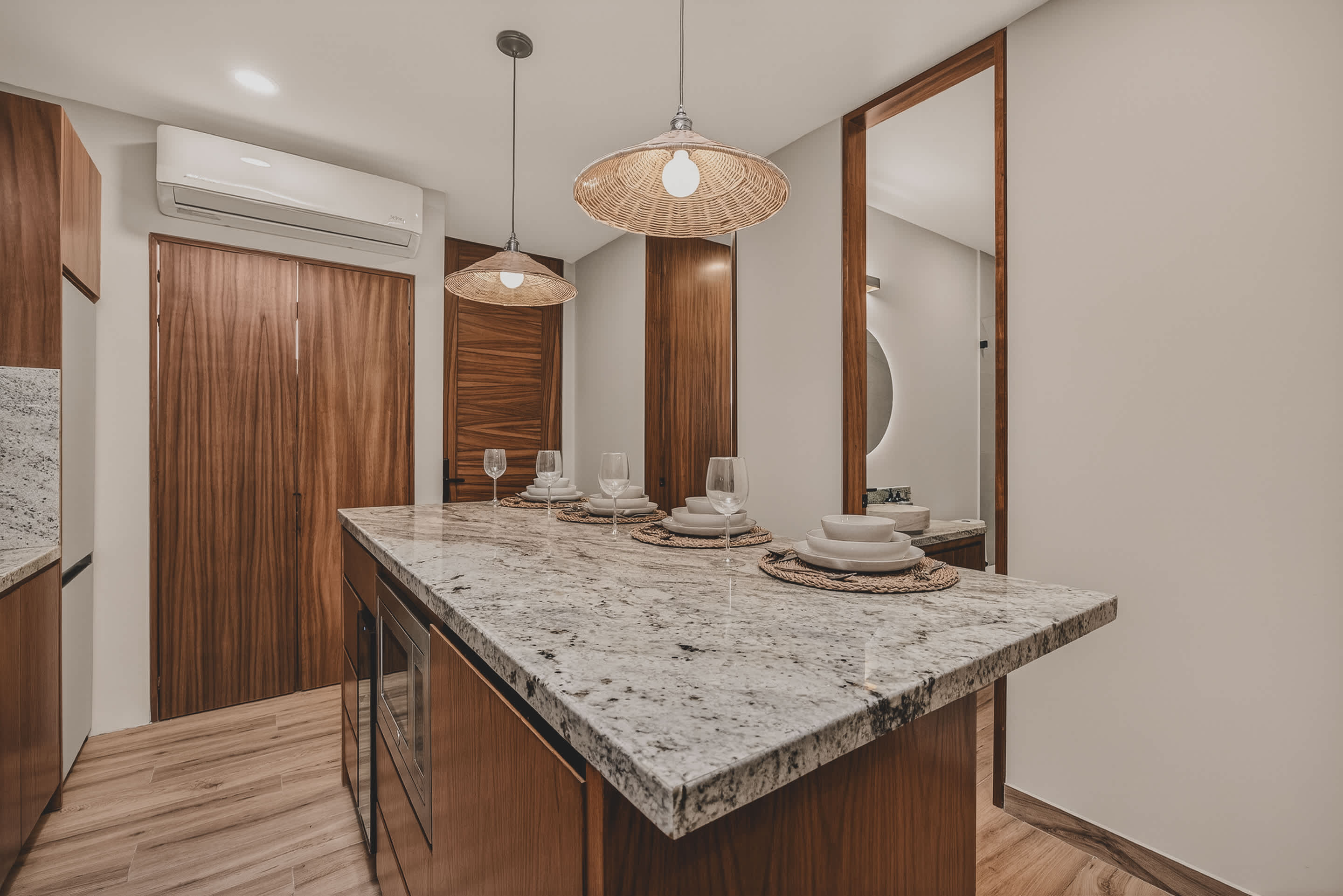 The kitchen island with granite countertop is set for dining, featuring woven placemats and wine glasses. Two woven pendant lights hang above, and a large mirror reflects the space, with wooden doors leading to other rooms.