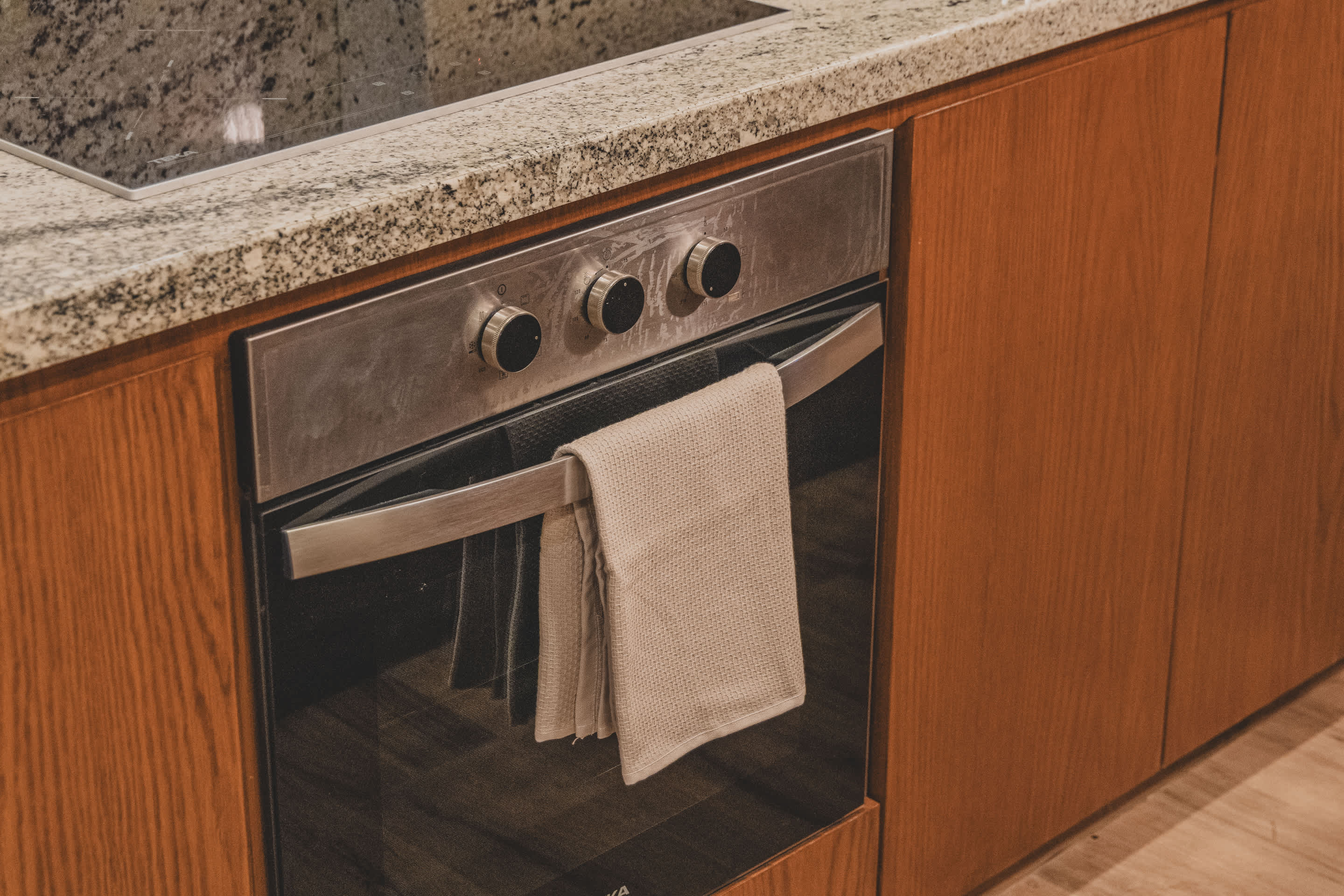 A stainless steel built-in oven with control knobs and a glass door, integrated into wooden kitchen cabinetry below a granite countertop. A light-colored towel hangs from its handle.