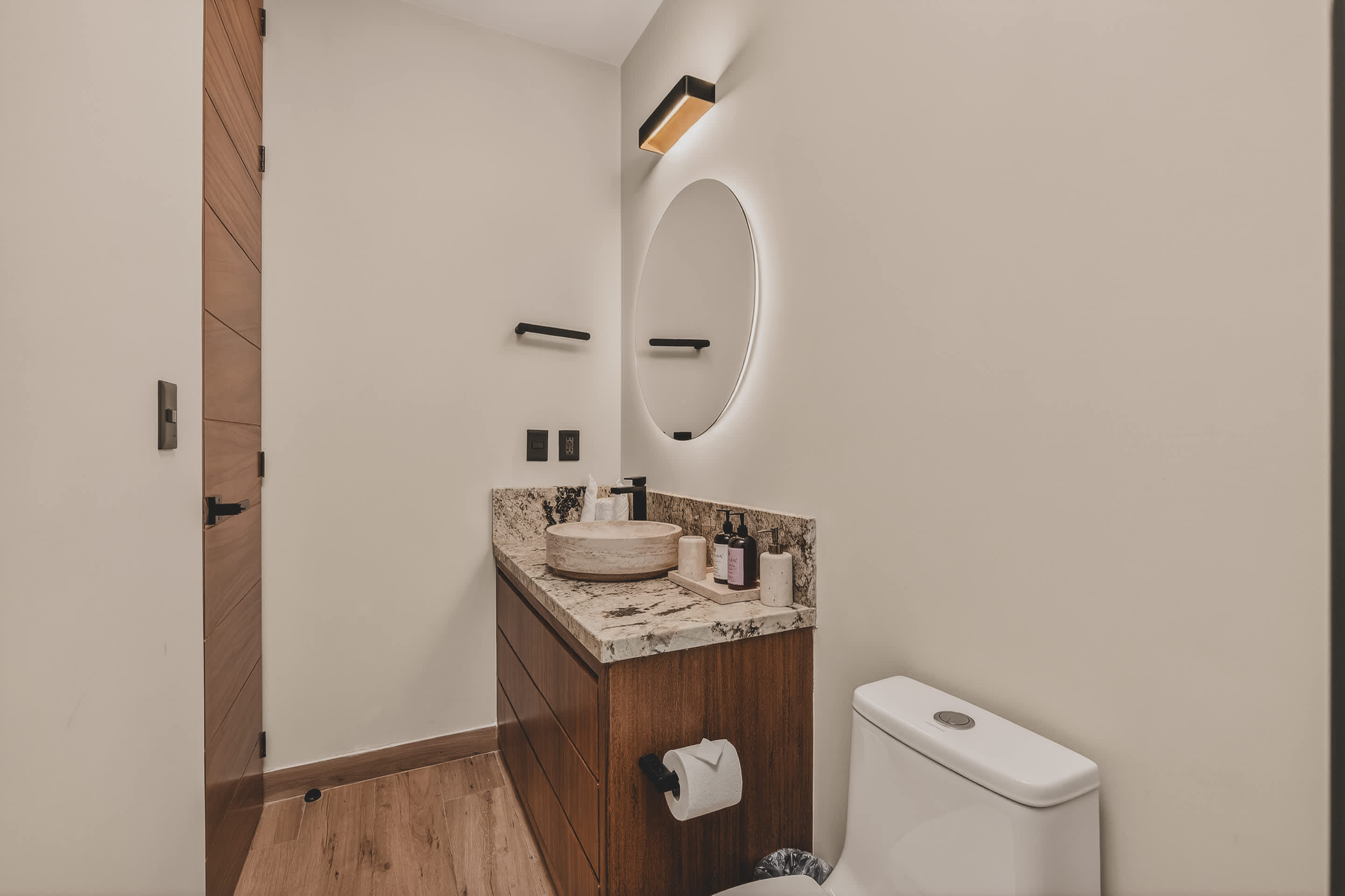This contemporary bathroom showcases a wooden vanity with a granite counter and vessel sink, a backlit circular mirror, and a white toilet, with a sleek wooden door to the left.