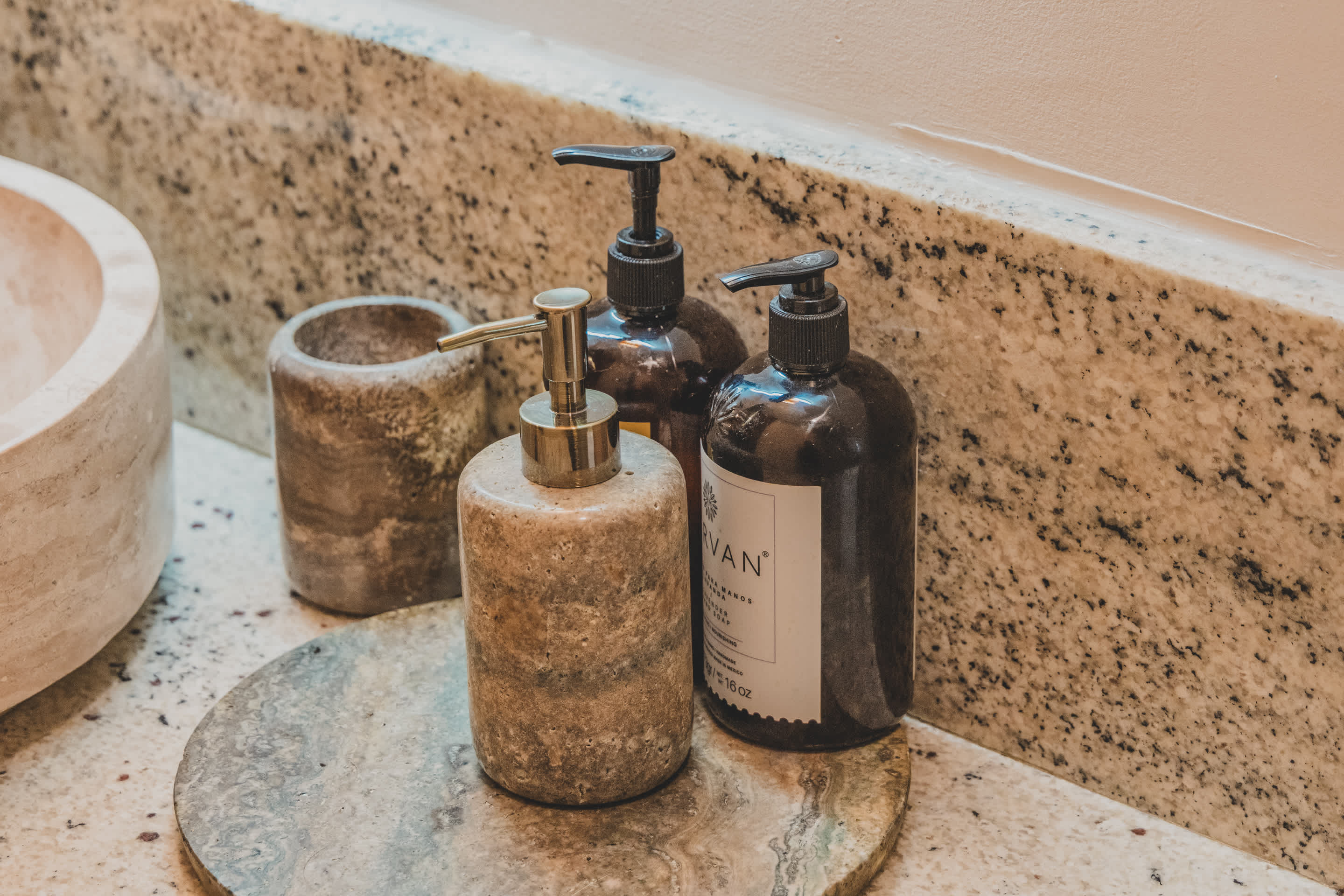 A close-up view of bathroom toiletries on a speckled granite countertop, including a stone pump dispenser and two dark pump bottles, arranged on a round stone tray next to a light-colored vessel sink.