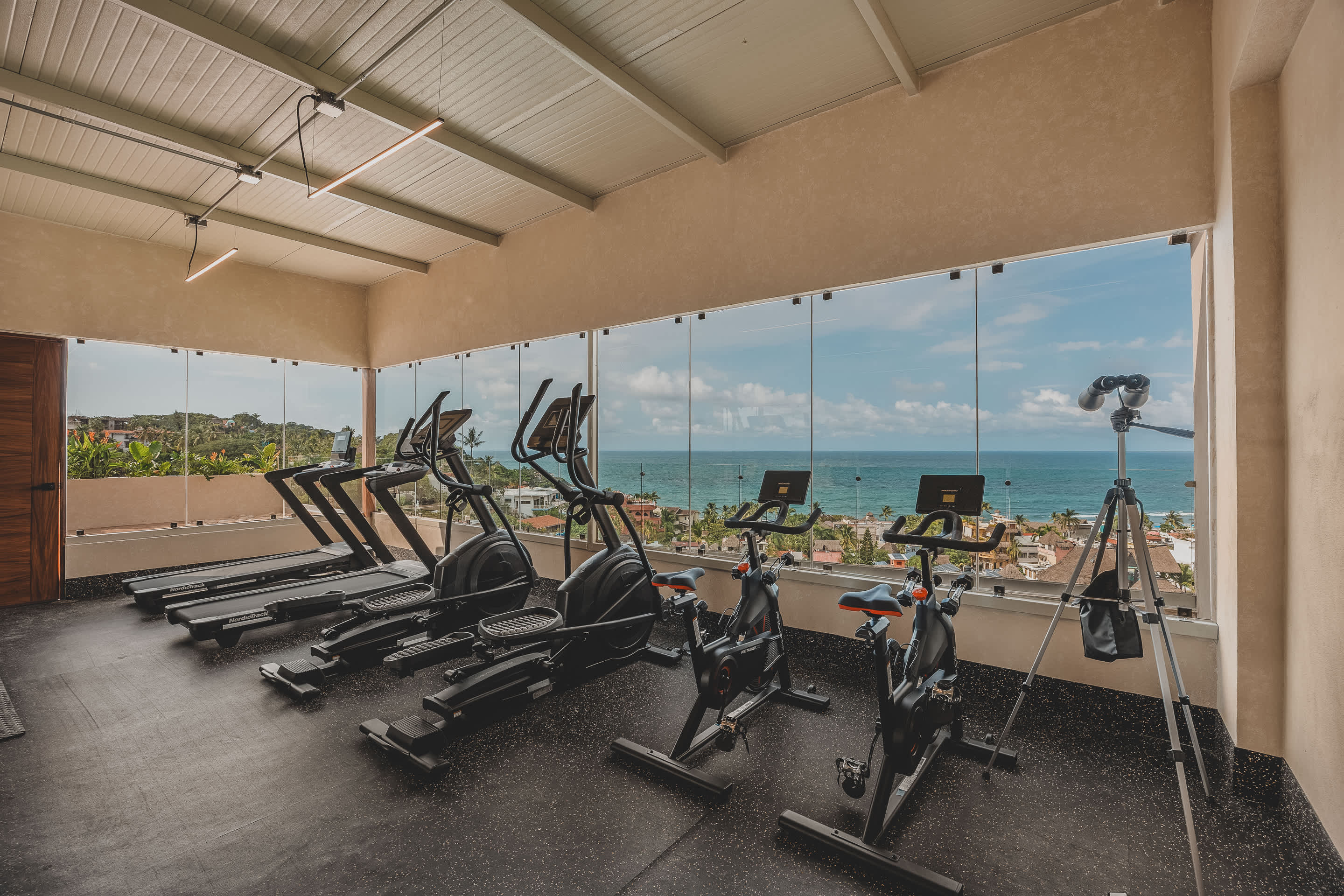 A well-equipped gym features several treadmills, ellipticals, and stationary bikes arranged to face large windows that offer a stunning panoramic view of the ocean and coastline.