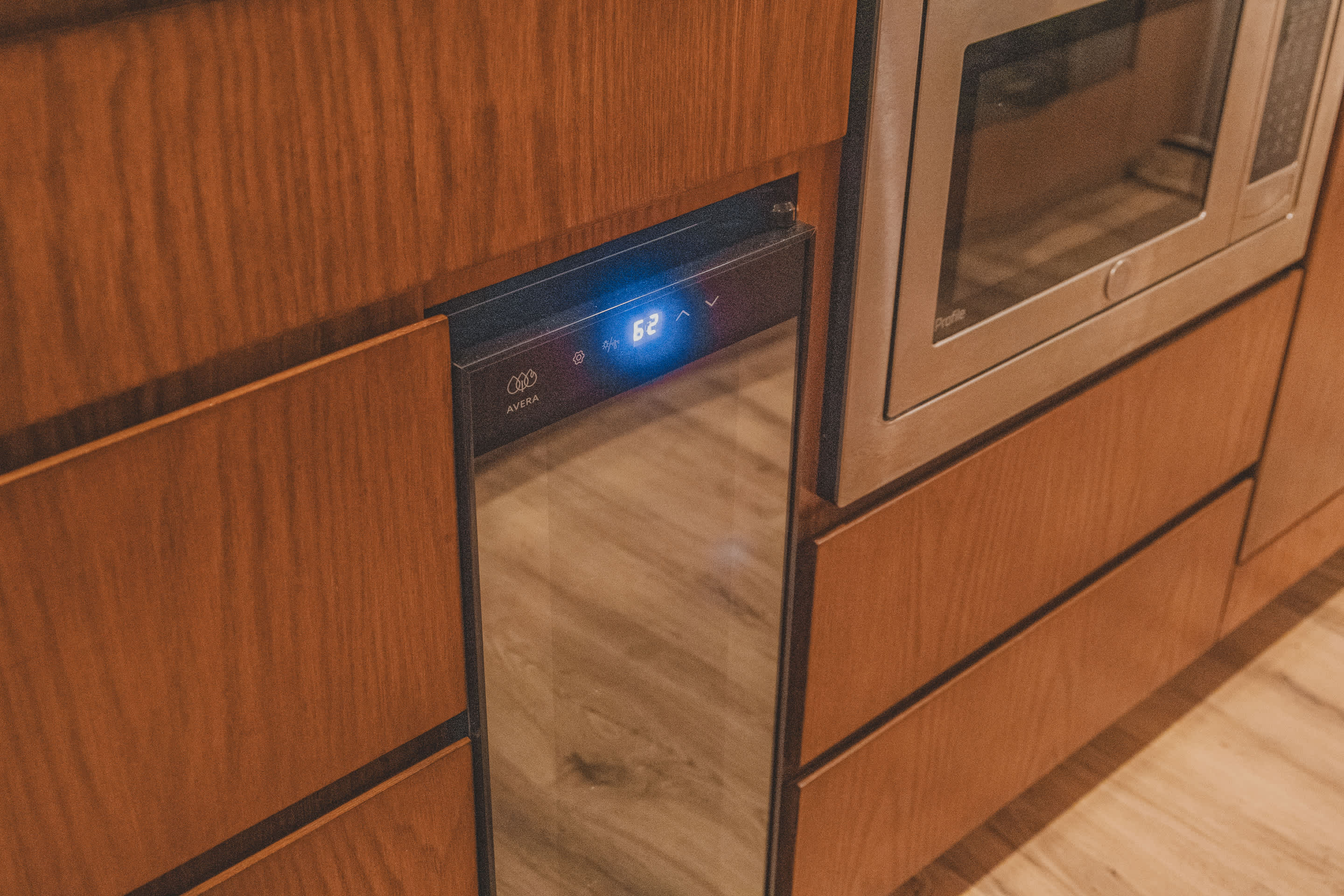 A built-in Avera wine cooler with a glass door and digital display showing '62' is integrated into wooden kitchen cabinetry, next to a stainless steel microwave.