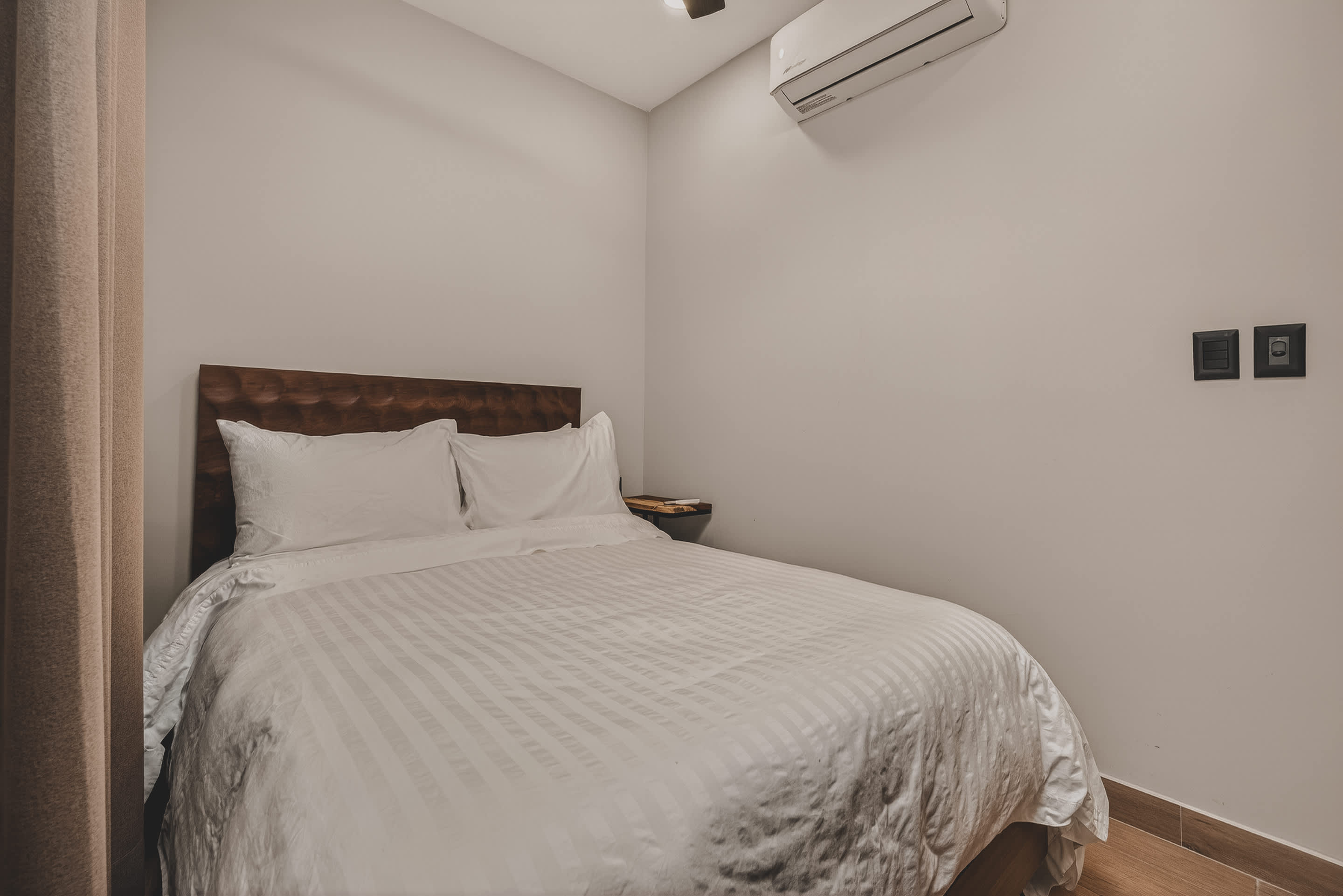 A minimalist bedroom with a white-striped queen bed and a dark wooden headboard against a light grey wall. An air conditioning unit is mounted above the bed, and a small bedside table is visible.