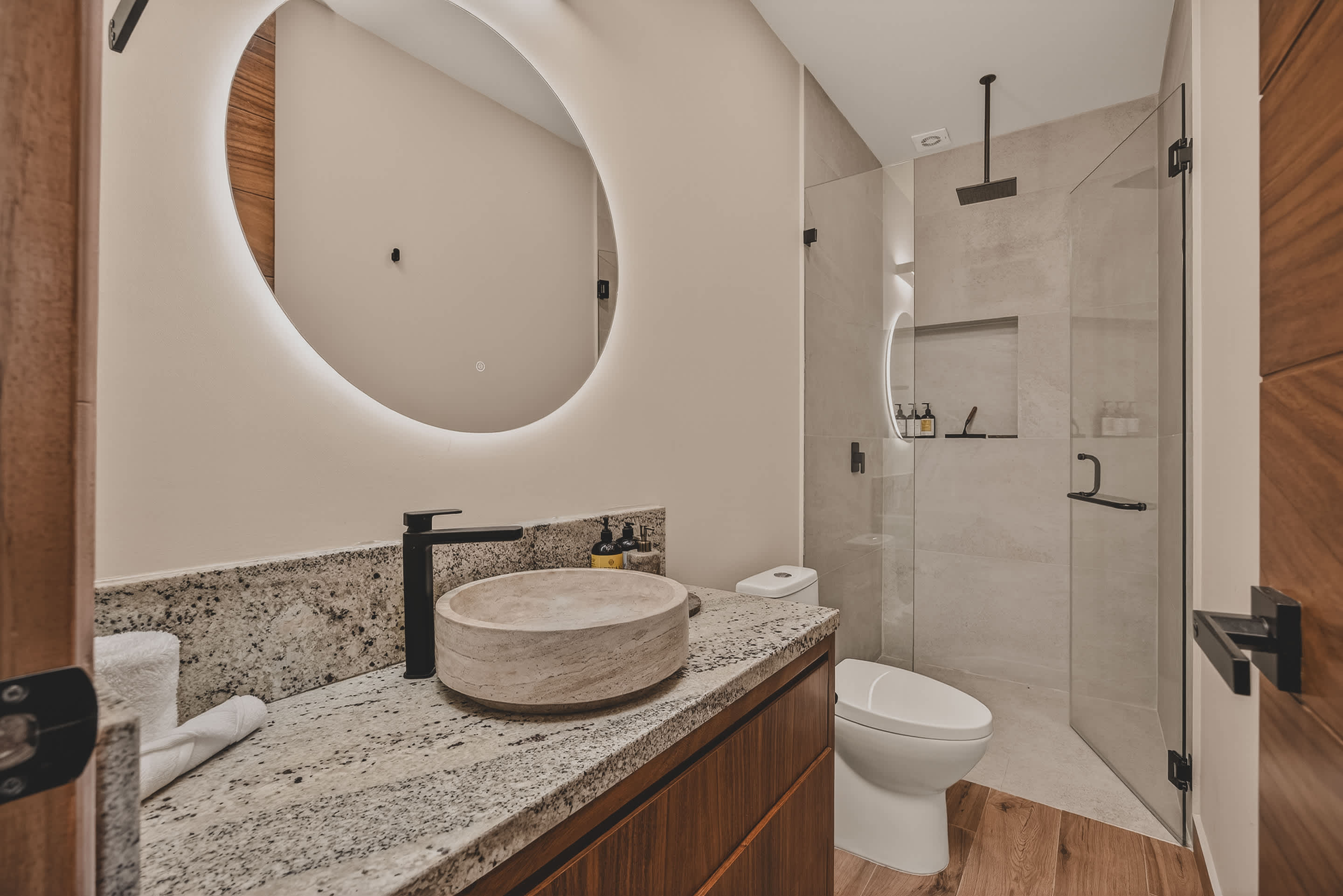 A bright bathroom features a wooden vanity with a granite top and stone vessel sink, a modern toilet, and a spacious walk-in shower with a rain showerhead and glass enclosure.