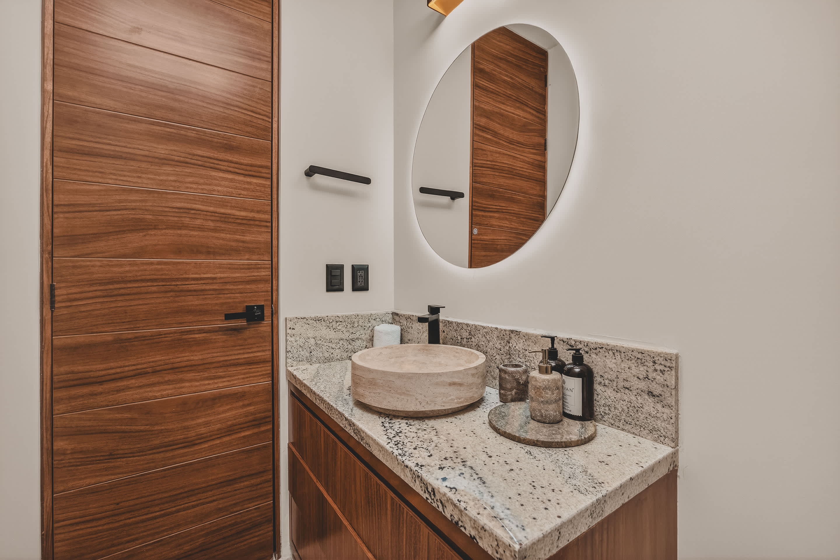 The bathroom vanity features a unique stone vessel sink on a granite countertop, adorned with modern black fixtures and a selection of toiletries, reflected in a large backlit circular mirror.
