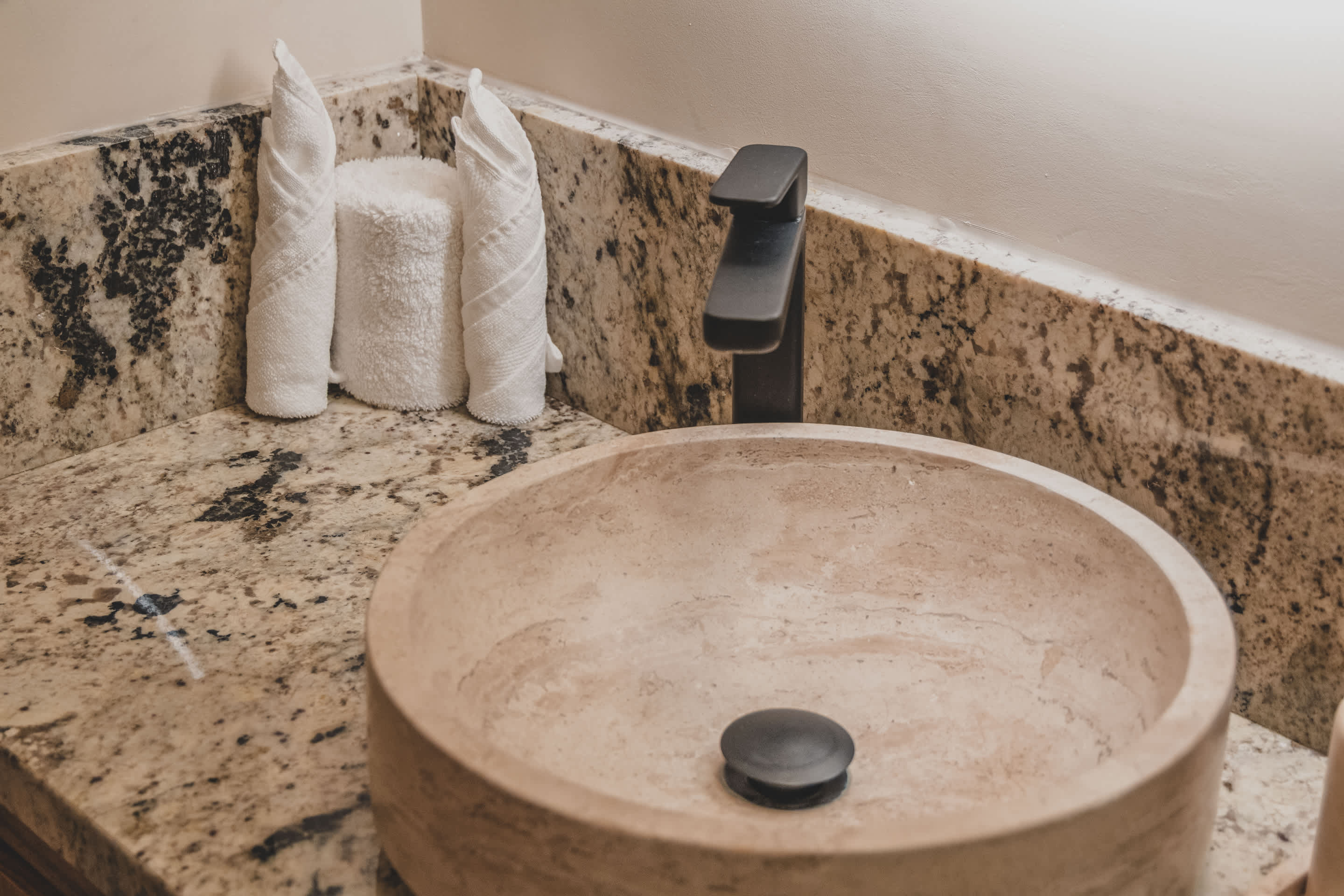 A stylish bathroom vanity showcases a round, light-colored stone vessel sink with a sleek black faucet, resting on a speckled granite countertop. Two white towels are artfully folded next to the sink.