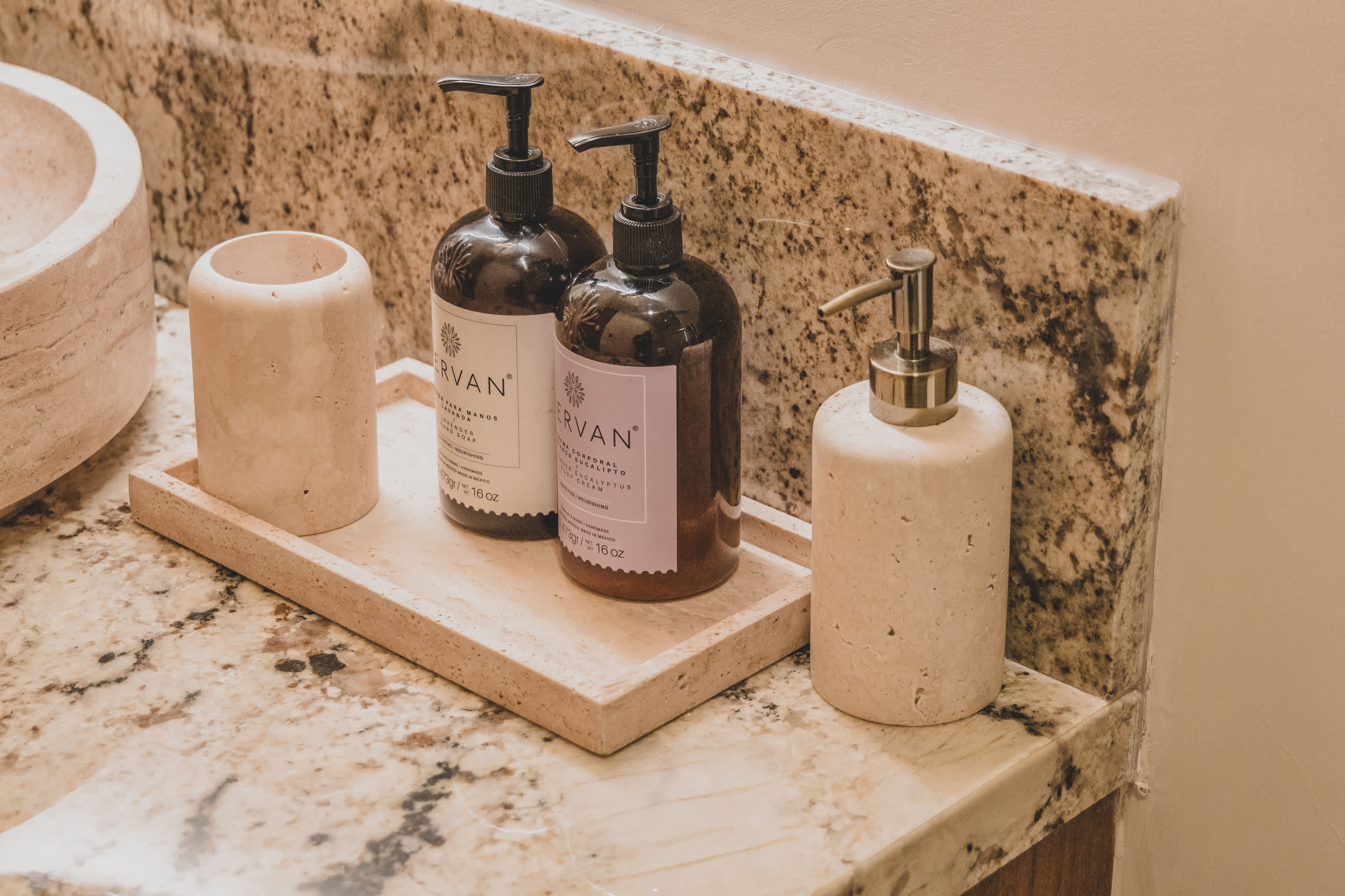 A collection of bathroom toiletries, including a light-colored stone pump dispenser and two dark pump bottles, are neatly arranged on a rectangular stone tray atop a speckled granite countertop.