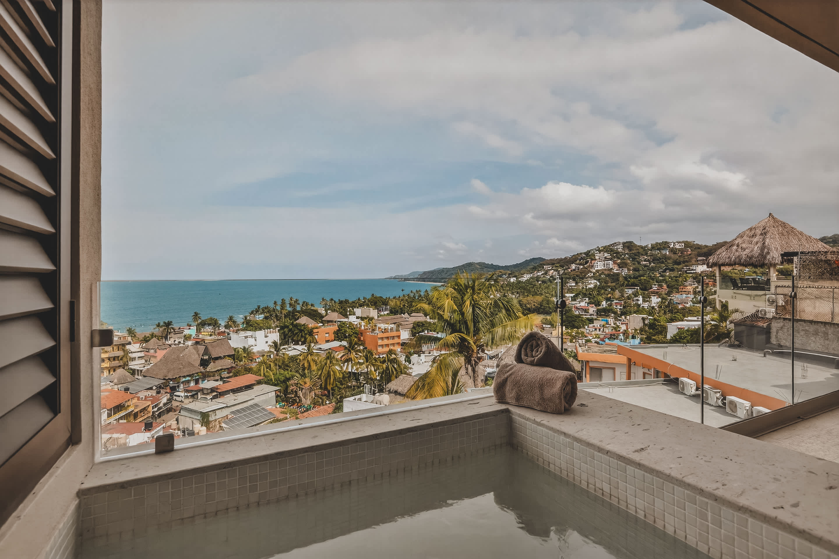 A private balcony hot tub with two rolled towels offers a stunning panoramic view of a vibrant coastal town, the vast ocean, and distant green hills under a cloudy sky.