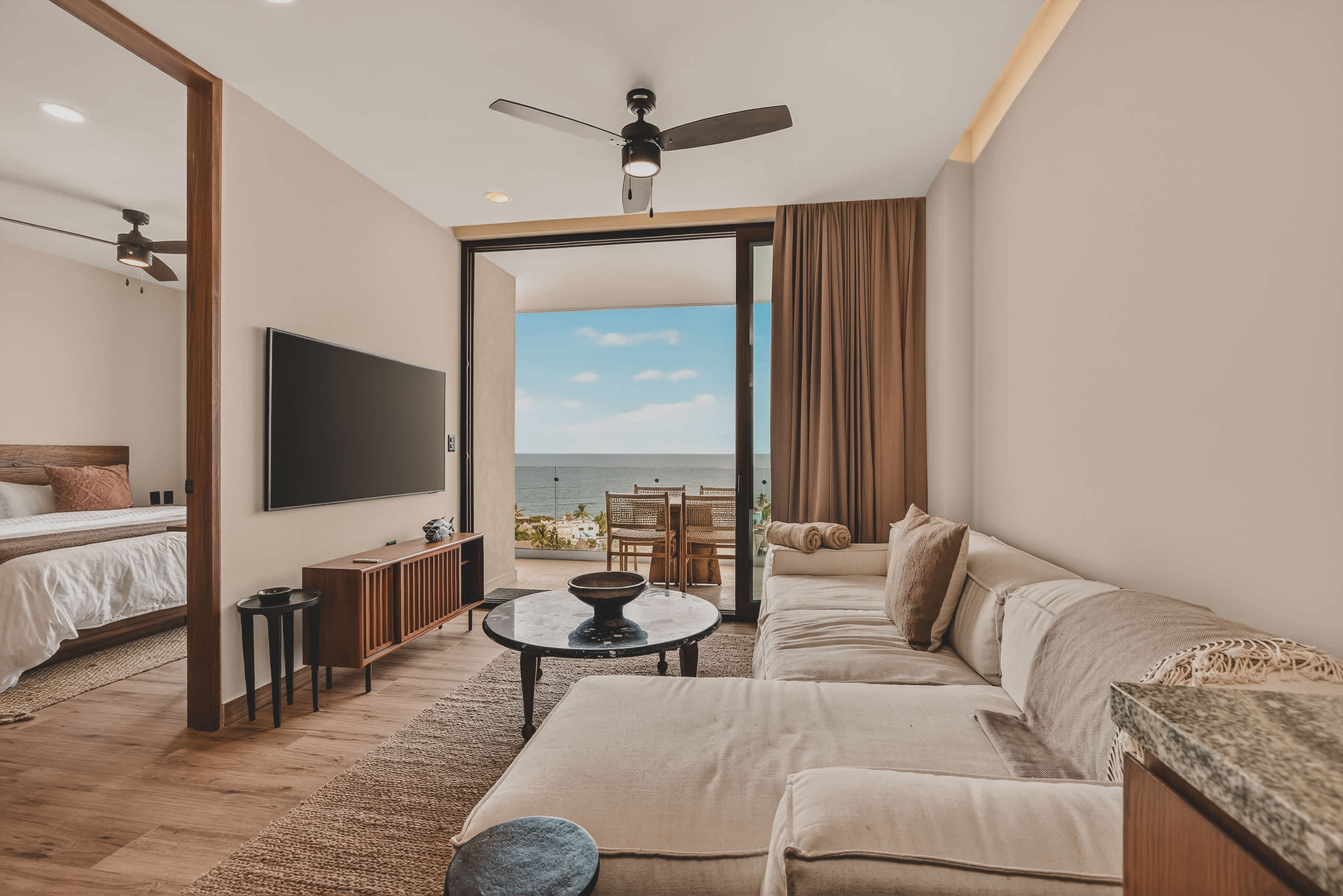 Contemporary living room featuring a white sectional sofa, a dark round coffee table, and a wall-mounted TV, with a partial view into an adjacent bedroom.