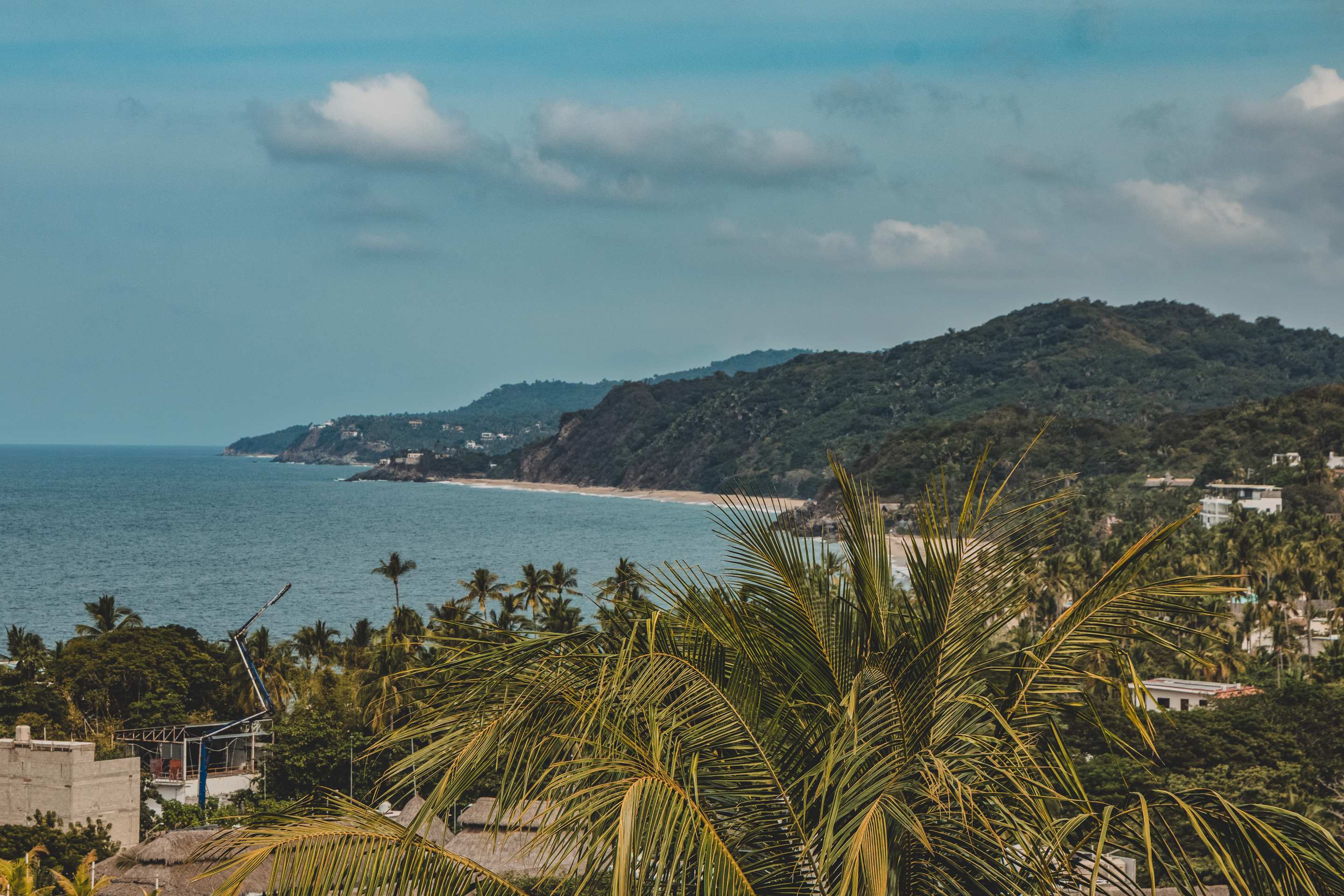 A scenic coastal view showcasing a long stretch of sandy beach bordered by lush green hills and the vast blue ocean under a partly cloudy sky. Palm trees frame the foreground, adding to the tropical ambiance.