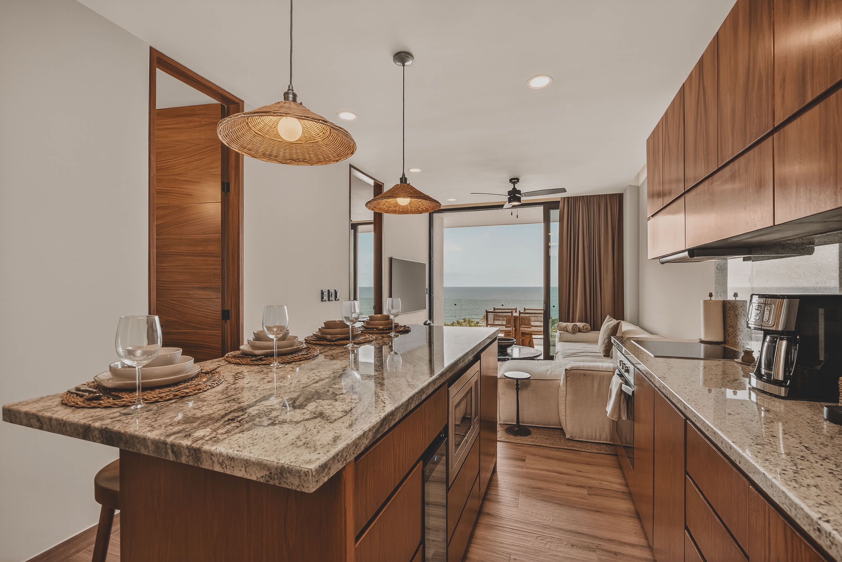 An open-concept living space featuring a kitchen island with dining settings, leading into a cozy living room with a white sectional sofa. Large sliding glass doors offer panoramic ocean views, connecting to an outdoor area.