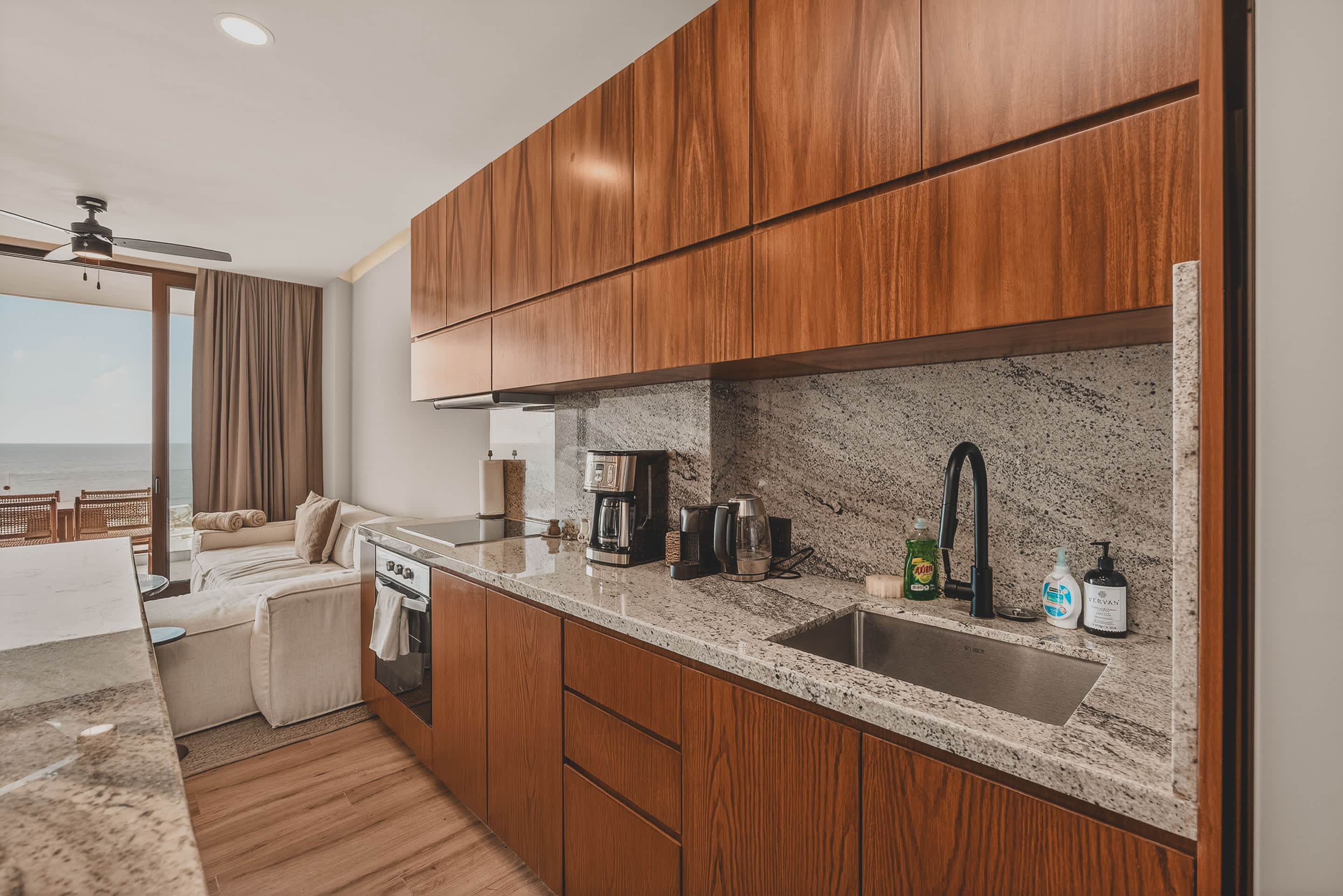A modern kitchen with wooden cabinets and granite countertops, featuring a stainless steel sink and various kitchen essentials. The open-concept design allows a clear view into the living area and out to the ocean through large glass doors.