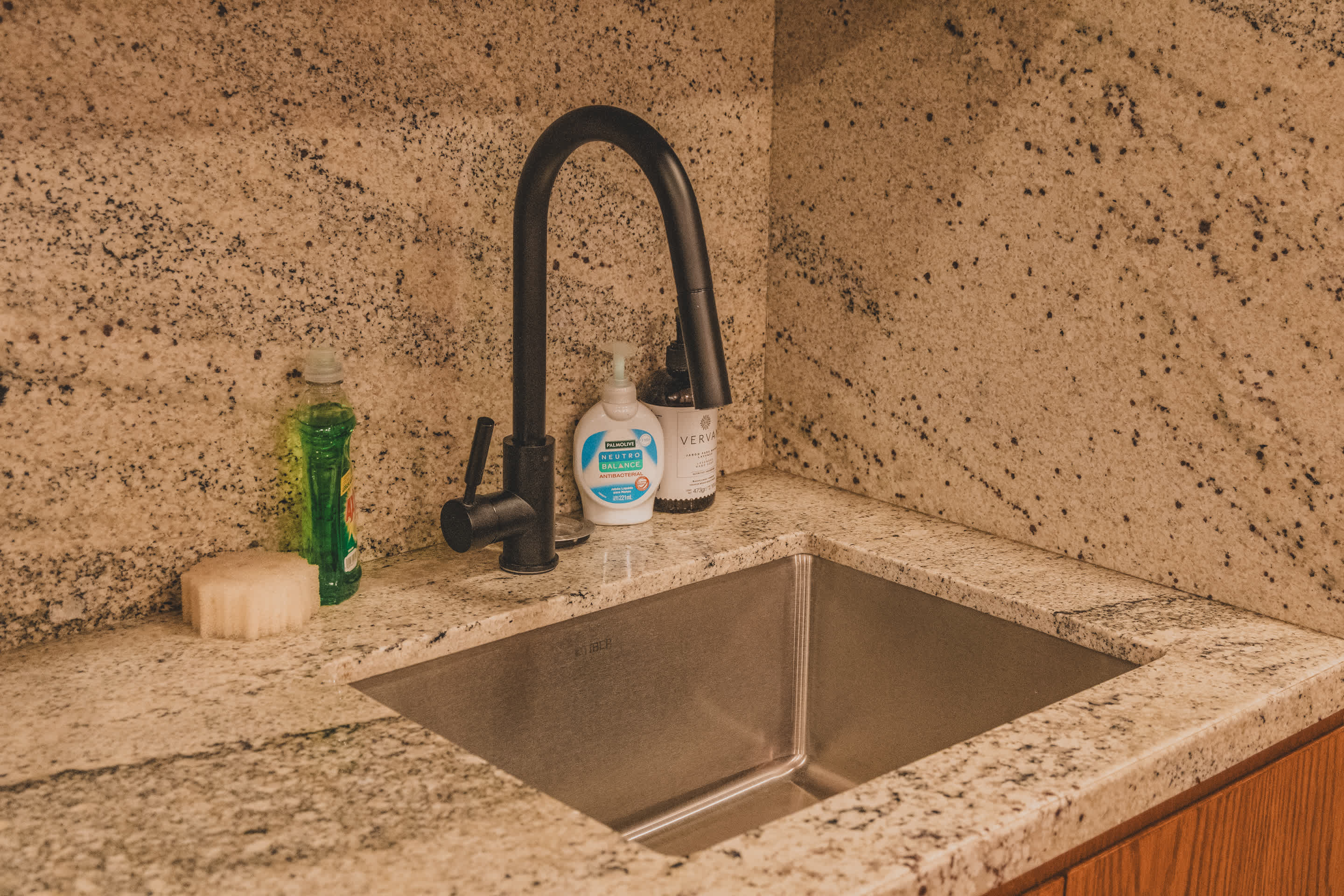 A modern square stainless steel sink with a black gooseneck faucet, set into a speckled granite countertop against a matching granite backsplash. Various soap dispensers are visible.