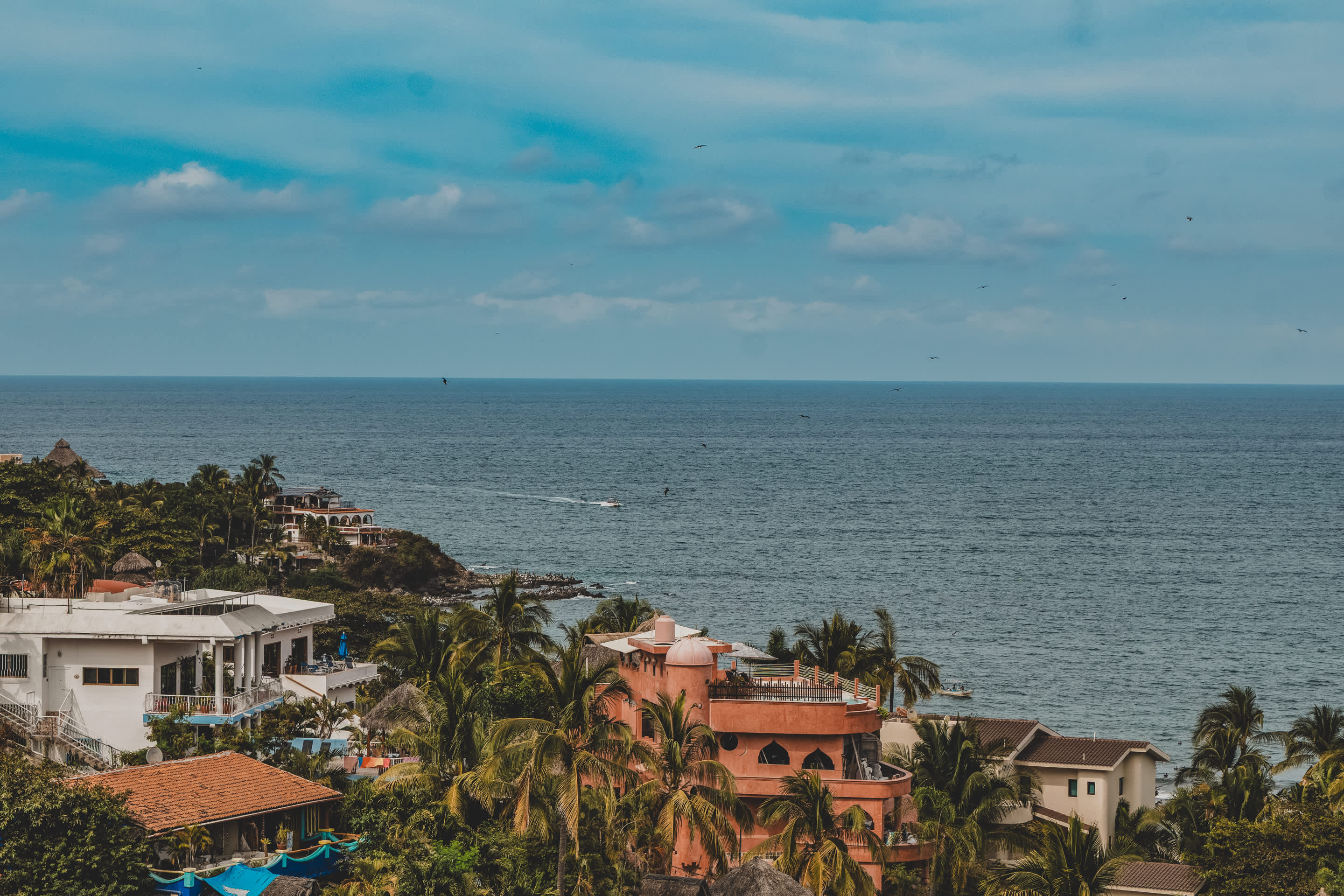 A panoramic ocean view captures the vast blue sea meeting a lush coastline dotted with white and terracotta buildings and abundant palm trees. Several boats are visible on the water under a partly cloudy sky.