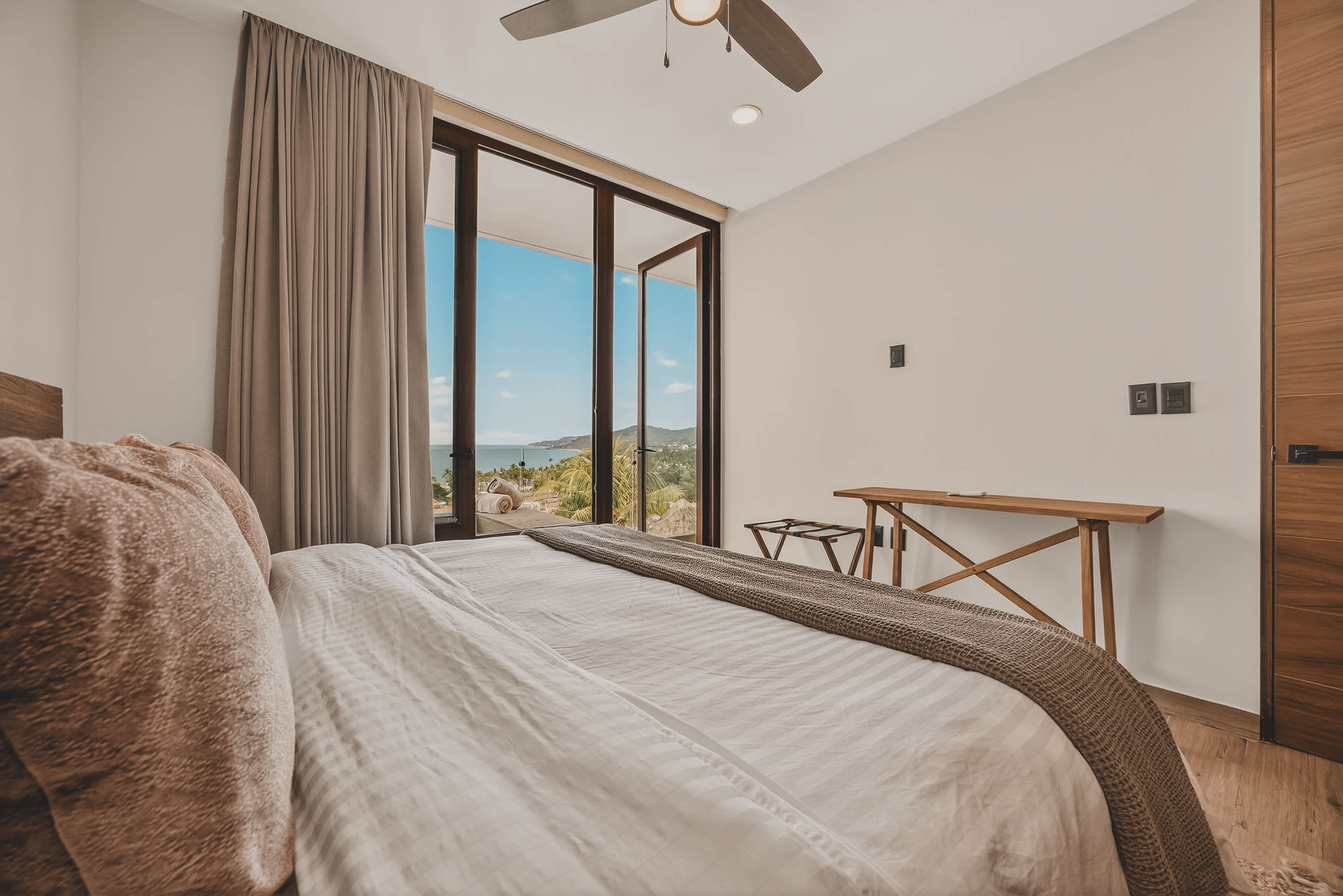 A bright bedroom with a large bed featuring white and brown bedding, facing expansive glass doors that open to an ocean view with lush greenery. A ceiling fan and a wooden side table with a luggage rack are visible.