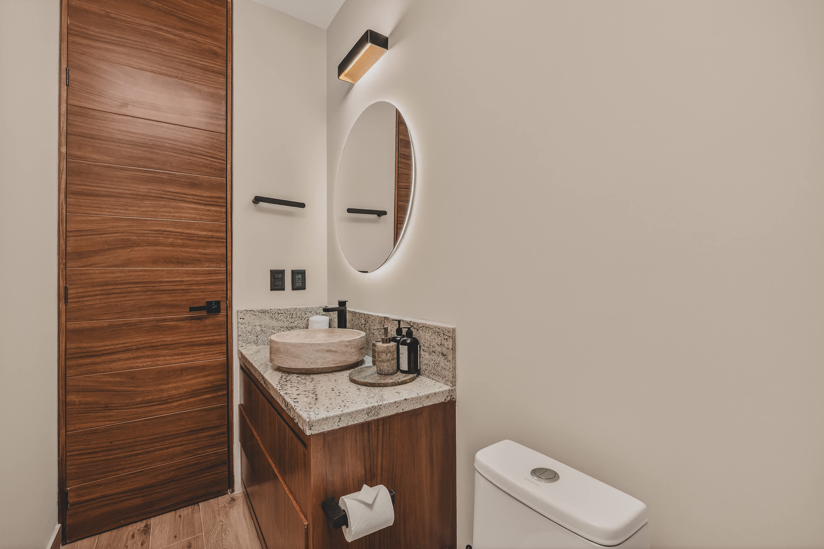 A modern bathroom featuring a light-colored, circular vessel sink on a granite countertop, a black wall-mounted faucet, and a large round mirror with backlighting. A white toilet is visible to the right.