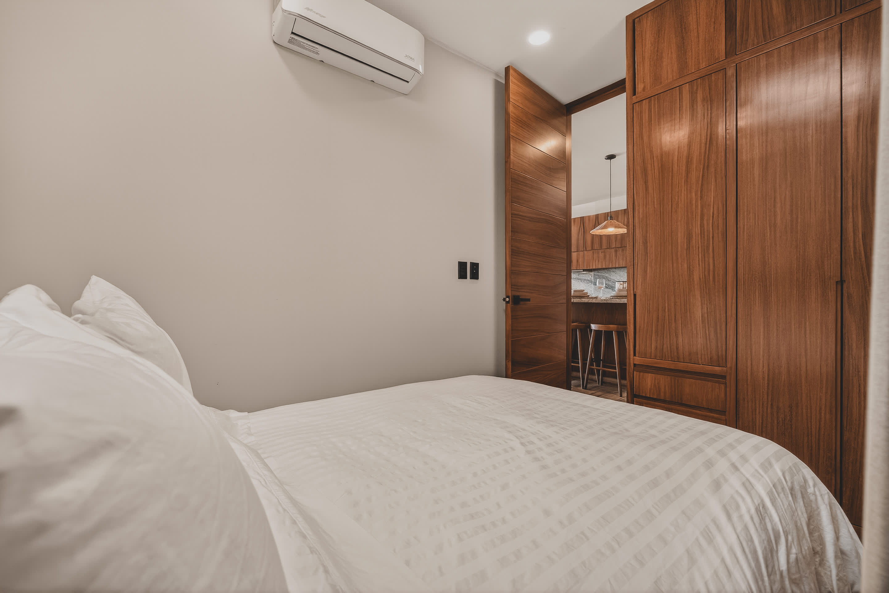 A bright bedroom featuring a white-striped queen bed with a dark wooden headboard, and extensive built-in wooden cabinetry. An open wooden door leads to a modern kitchen area with bar seating.