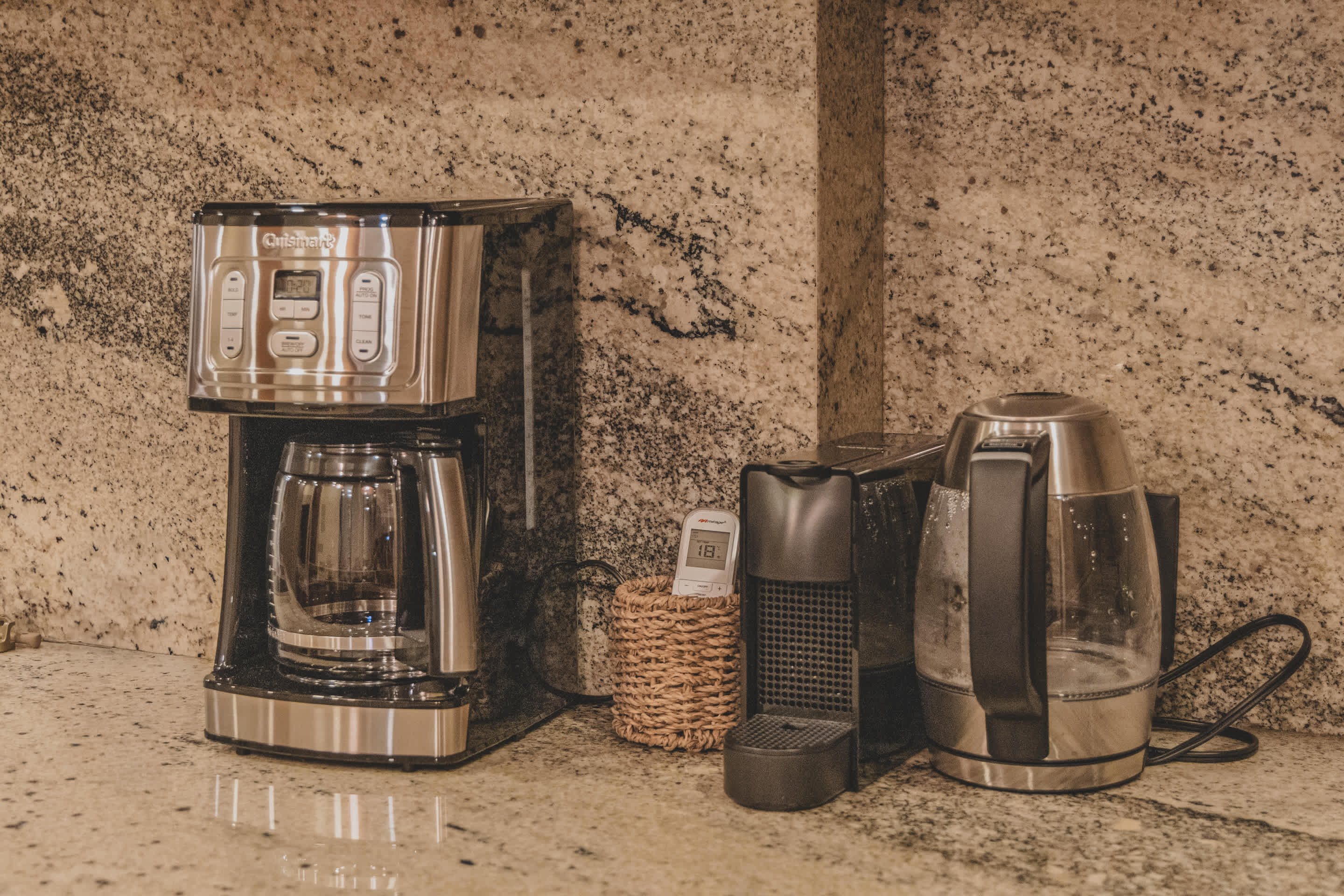 A coffee station on a speckled granite countertop, featuring a Cuisinart coffee maker, a Nespresso machine, and a modern electric kettle, all against a matching granite backsplash.