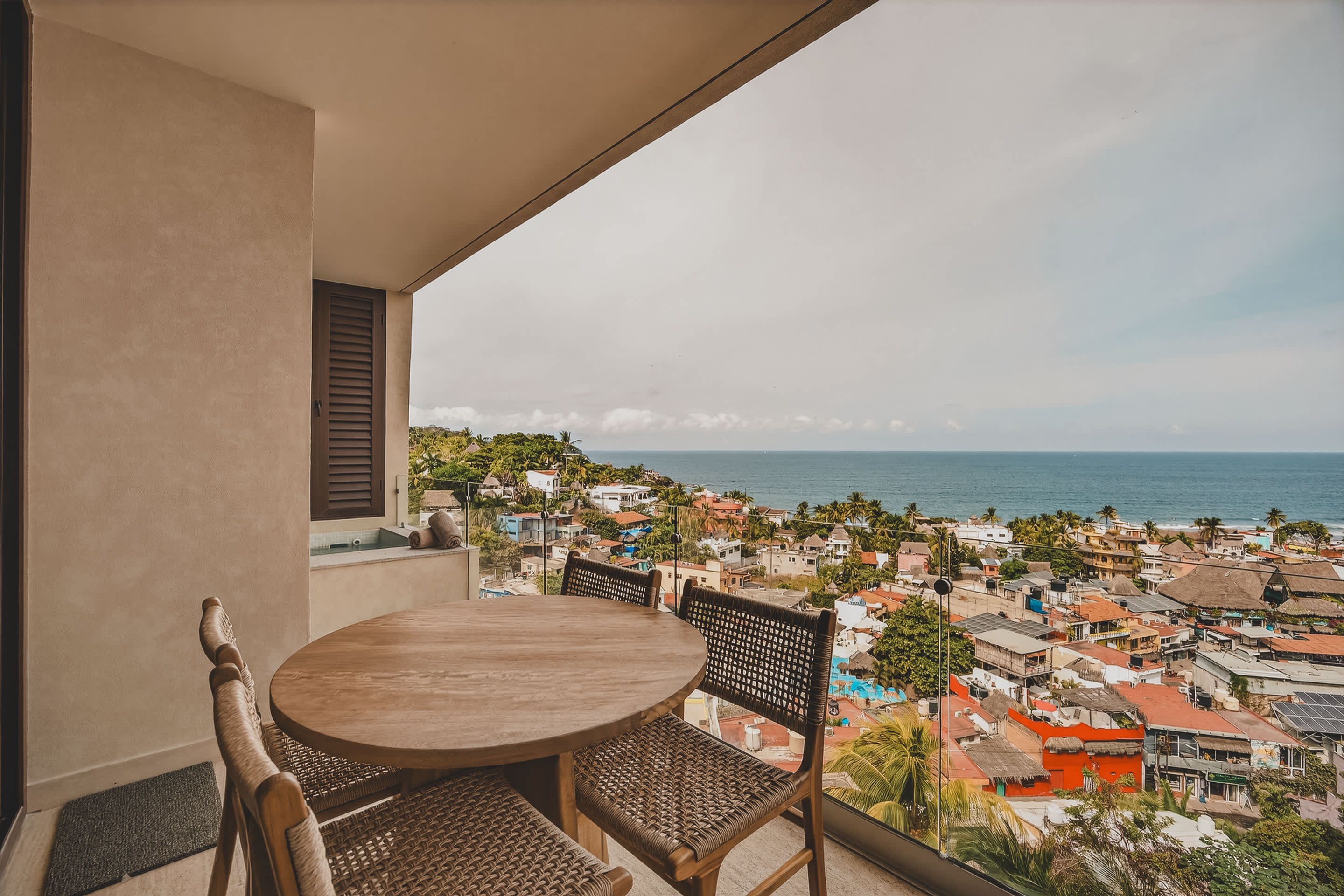 A spacious balcony features a round wooden dining table with four woven chairs, offering an expansive view of a bustling coastal town and the serene ocean under a bright sky.