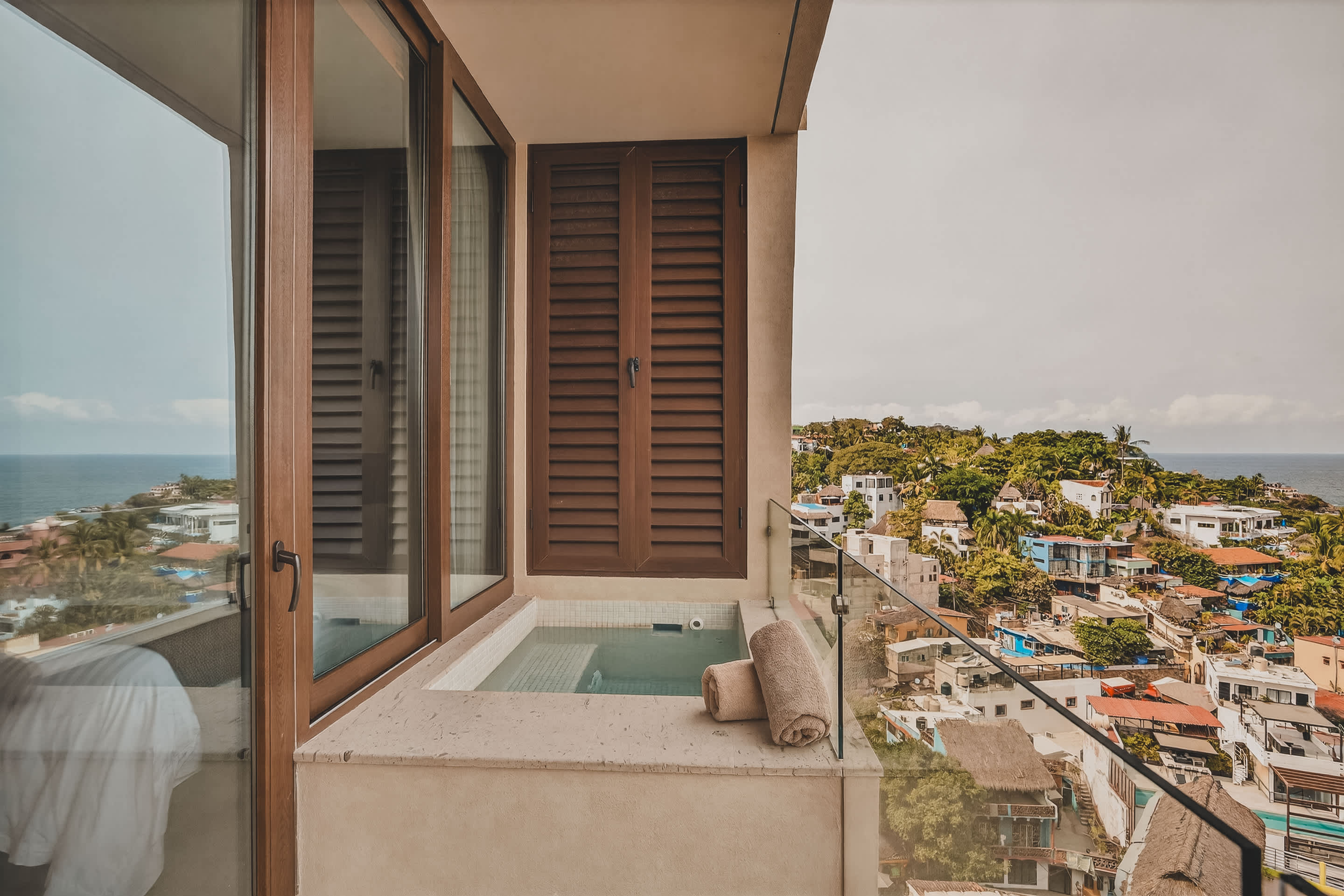 A private balcony featuring a small, square plunge pool or hot tub with rolled towels, enclosed by a glass railing. The balcony offers a scenic view of a bustling coastal town and the distant ocean.