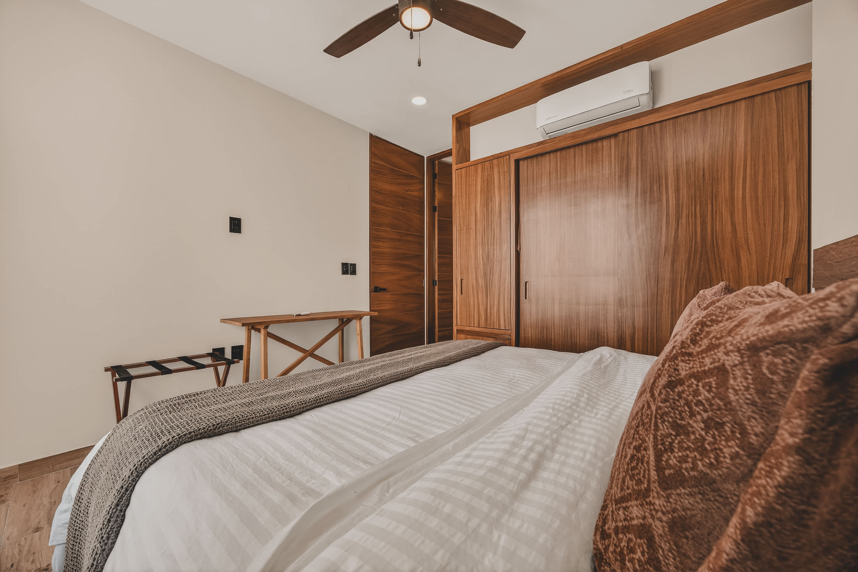 A modern bedroom with a large bed, featuring a wooden headboard and built-in wooden closets. An air conditioning unit is mounted above the closet, and a wooden desk with a luggage rack sits against the wall.