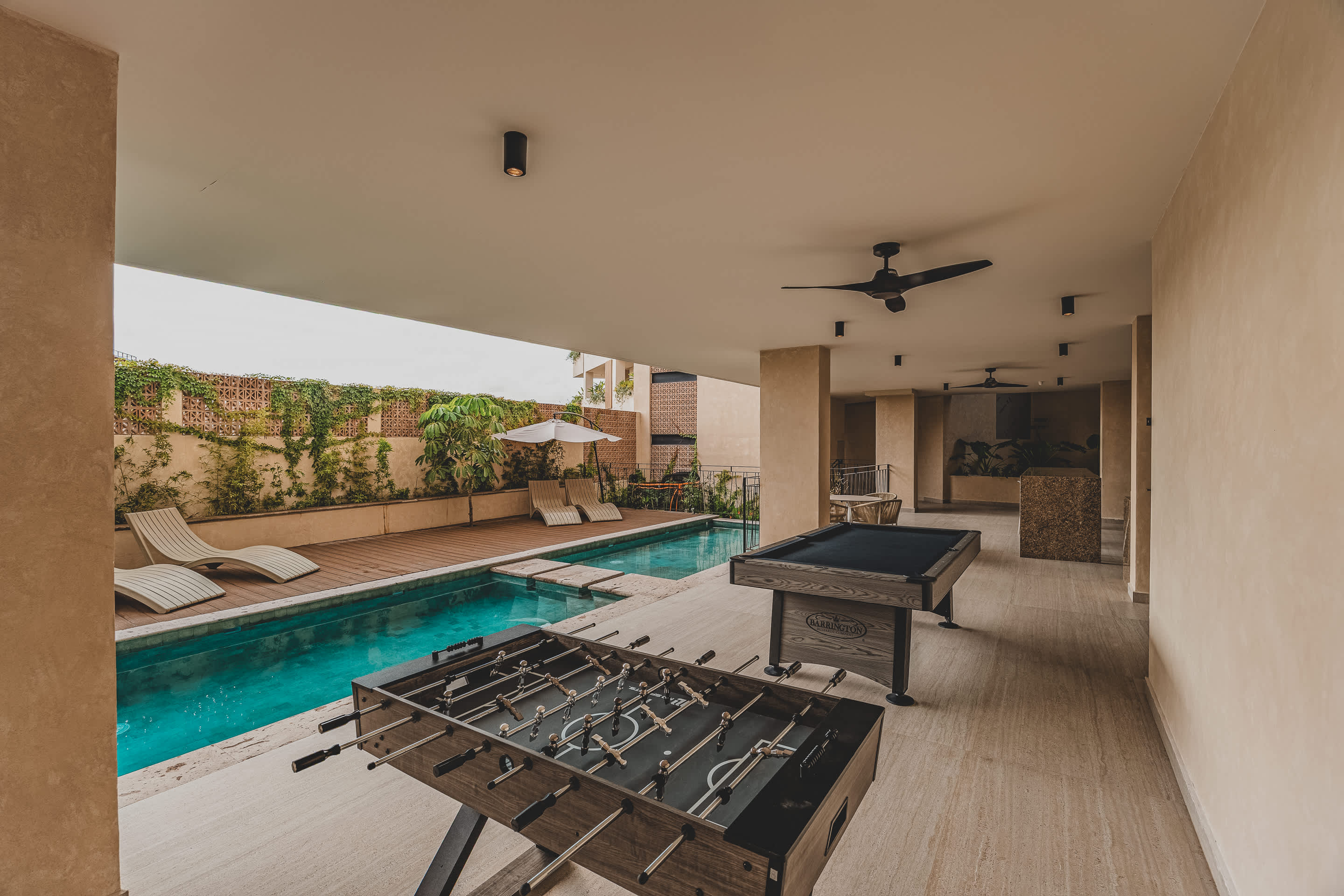 A spacious covered outdoor patio with a foosball table and a pool table, leading to a swimming pool area with lounge chairs and umbrellas. The flooring is light-colored tile, and the ceiling has recessed lighting and ceiling fans.