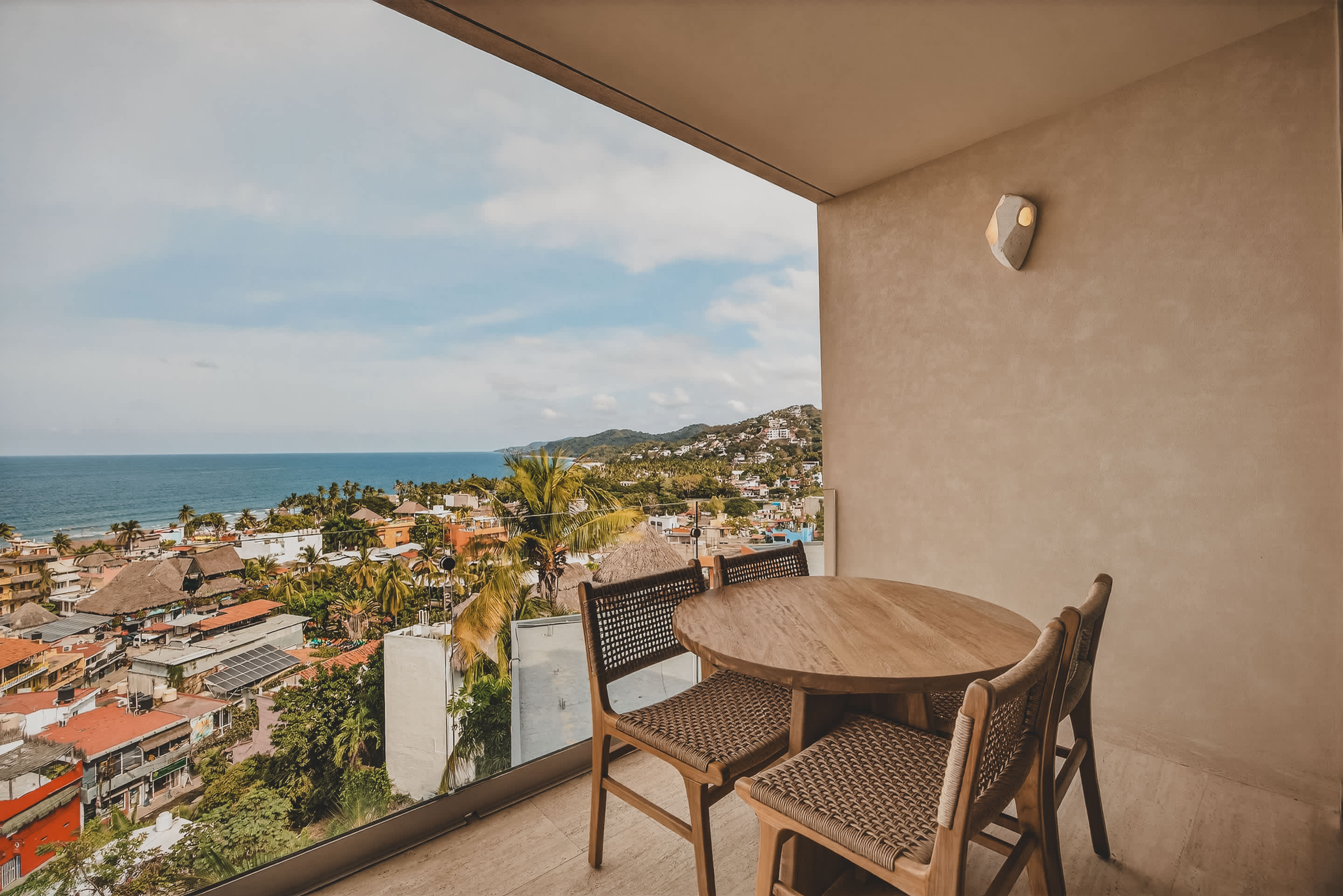 A cozy balcony dining area features a round wooden table and four woven chairs, set against a neutral wall, offering a sweeping vista of the coastal town and ocean.