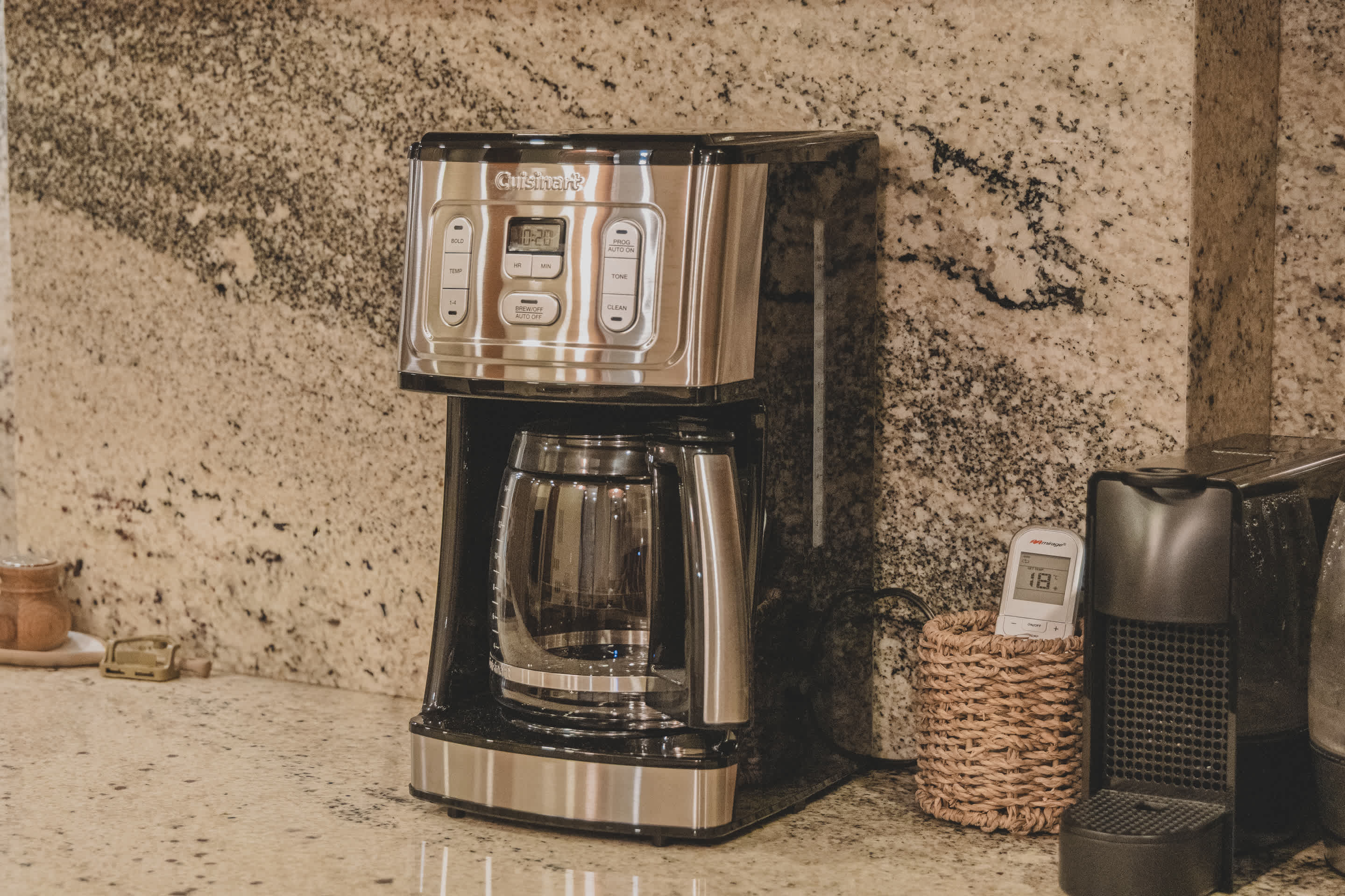 A Cuisinart coffee maker with a glass carafe sits on a speckled granite countertop against a matching granite backsplash, next to a small woven basket.
