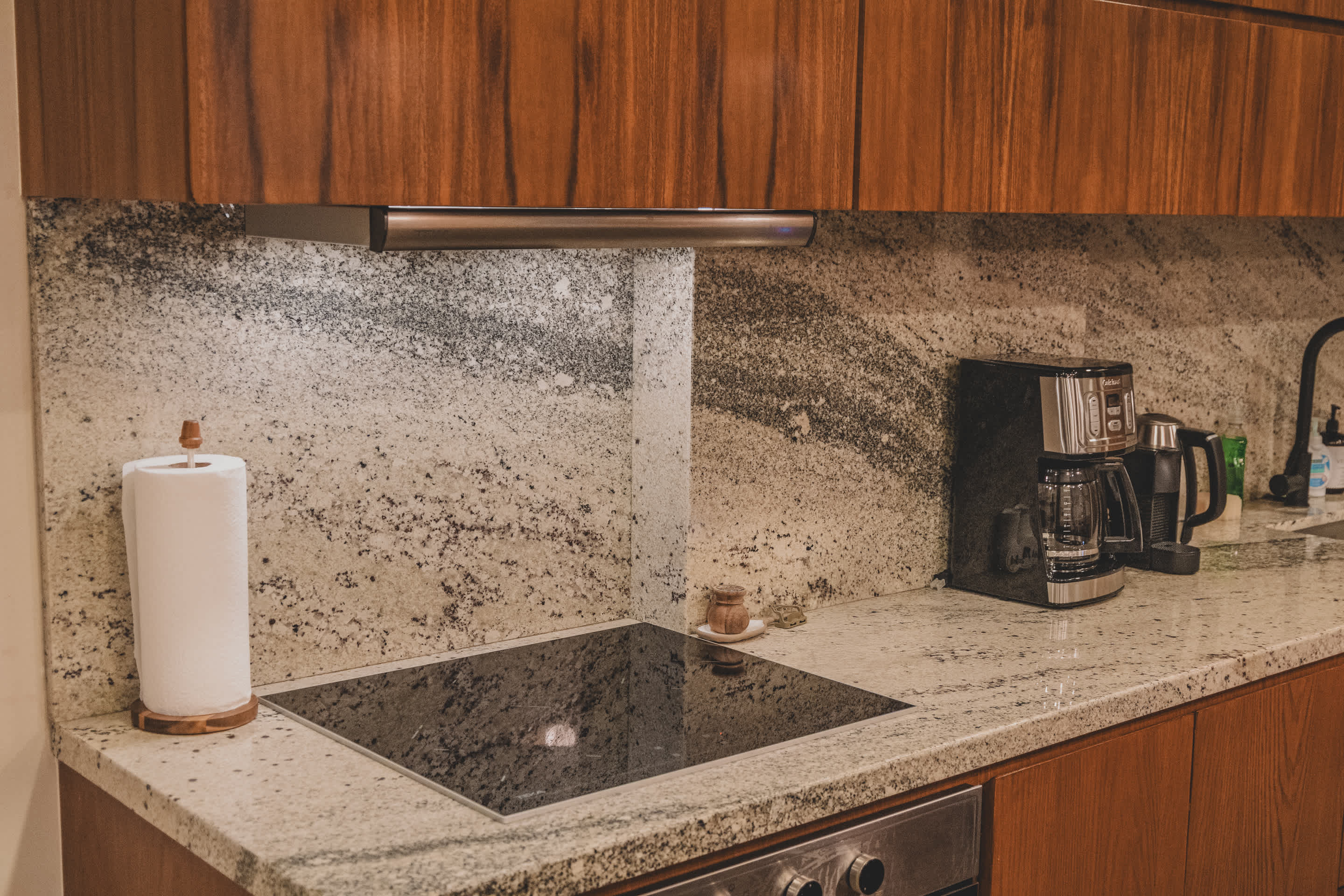 A contemporary kitchen features a black induction cooktop integrated into a speckled granite countertop, with matching granite backsplash. Wooden upper and lower cabinets frame the space, and a coffee maker and paper towel holder are visible.