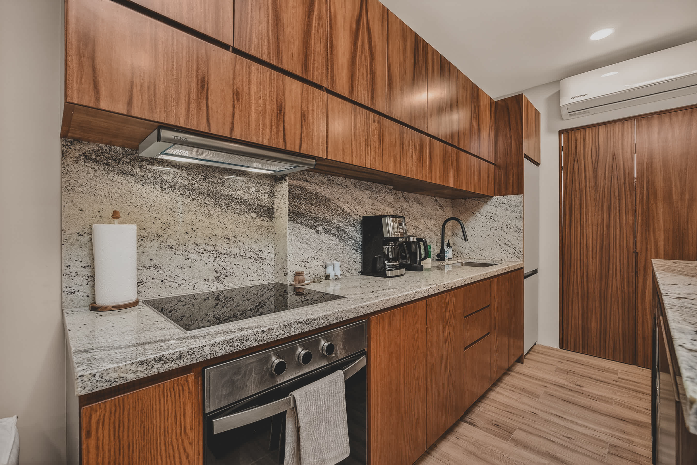 A detailed view of the kitchen counter showcasing light granite countertops, a built-in electric stovetop, and a stainless steel oven. Various kitchen appliances like a coffee maker and kettle are visible, along with wooden upper and lower cabinets.