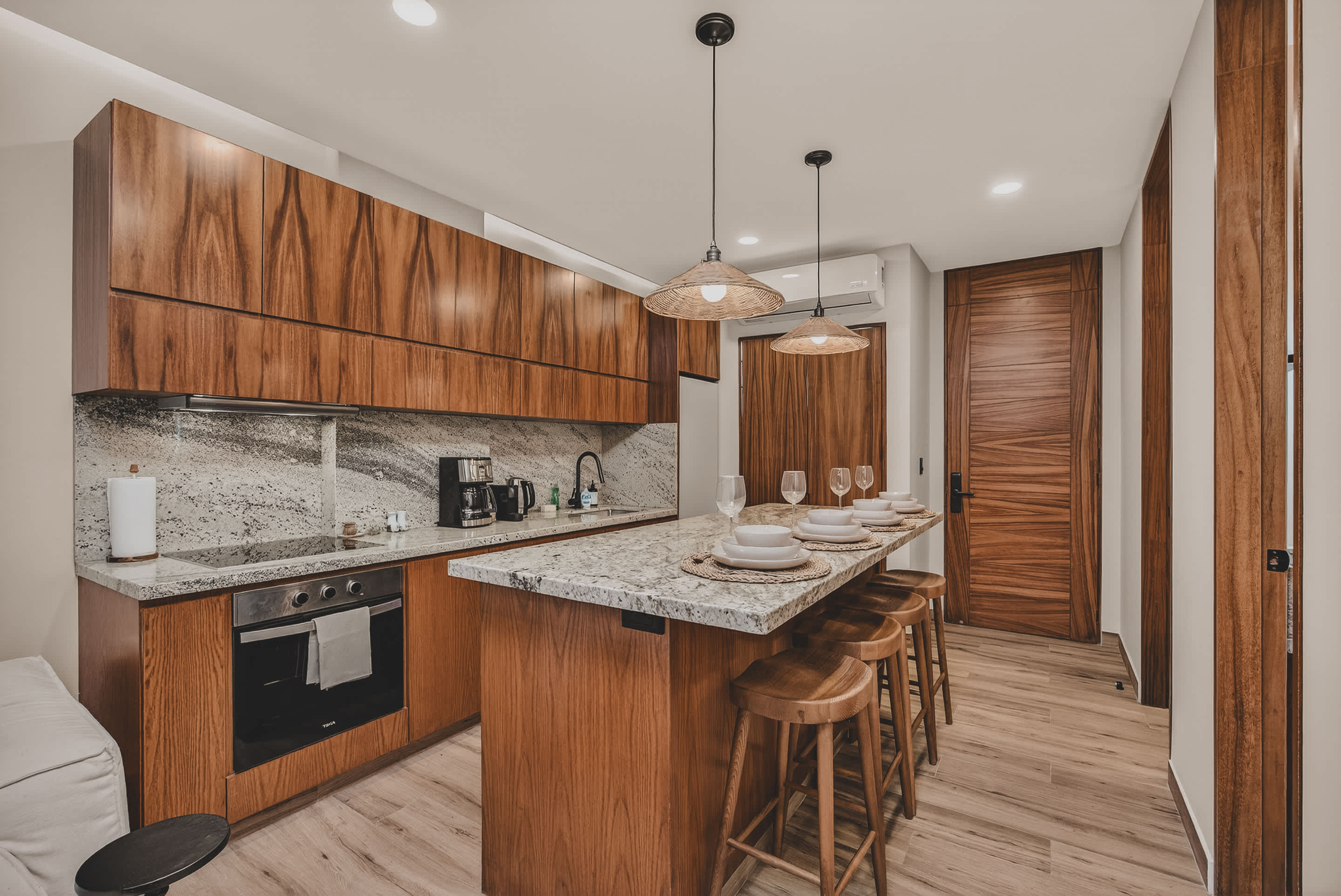 A modern kitchen featuring rich wooden cabinetry, light granite countertops, and a large island with seating for four. Integrated appliances include an oven, stovetop, and coffee maker, with woven pendant lights above the island.