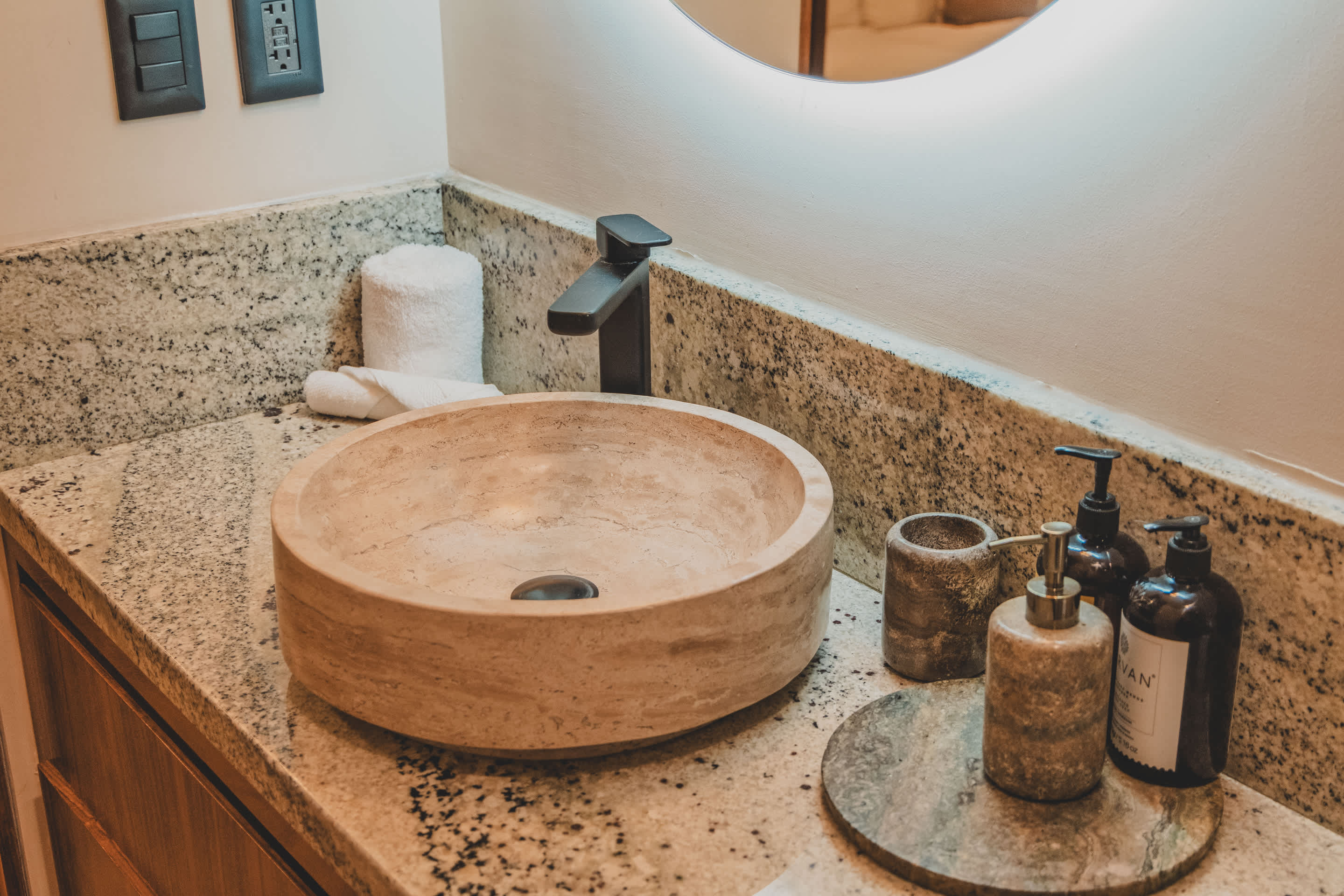 A modern bathroom vanity features a round, light-colored stone vessel sink with a matte black faucet set against a speckled granite countertop. Toiletries and a rolled white towel are neatly arranged beside the sink.