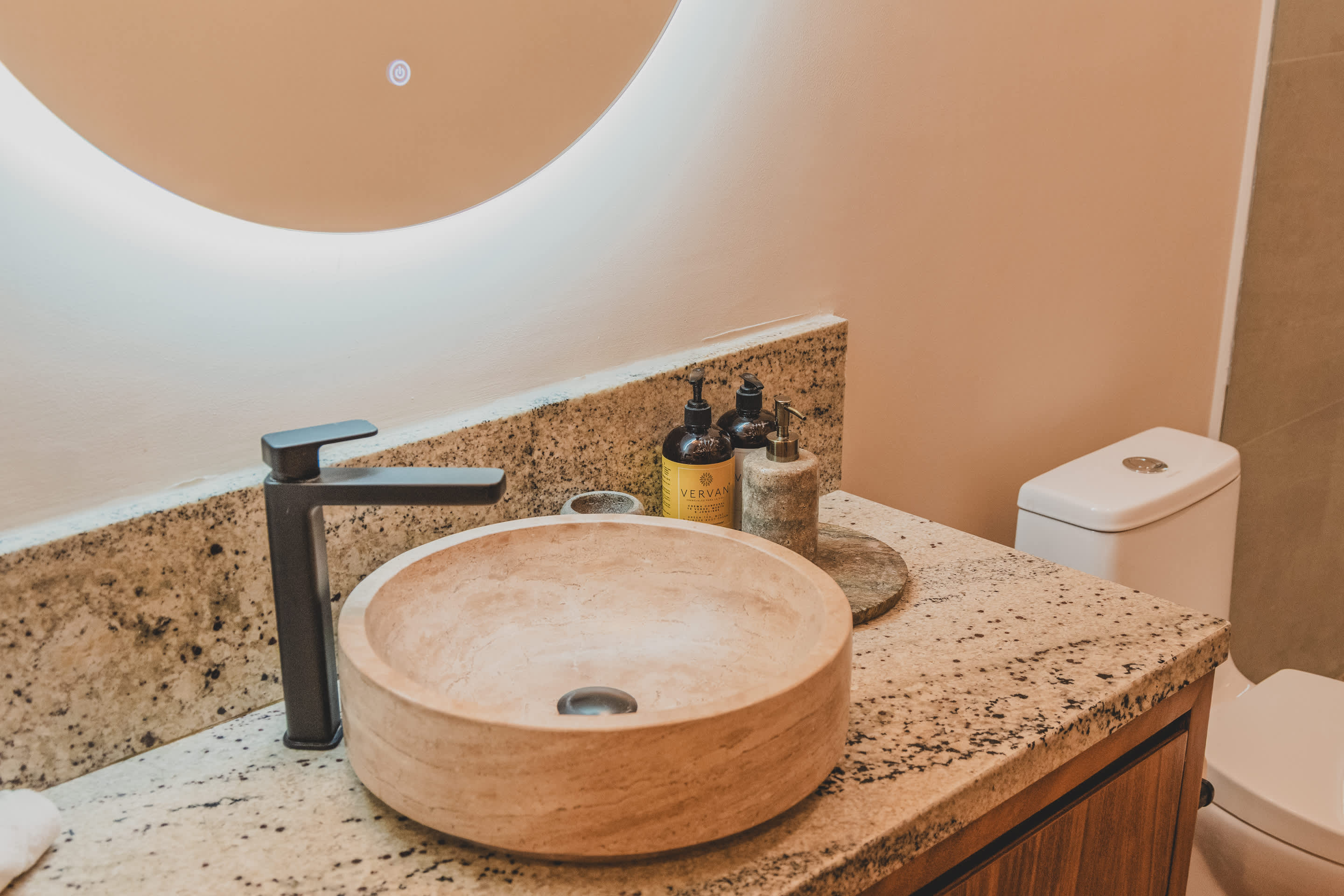 A close-up of a light-colored, circular vessel sink with a black wall-mounted faucet on a granite countertop, featuring various soap bottles and a backlit round mirror.