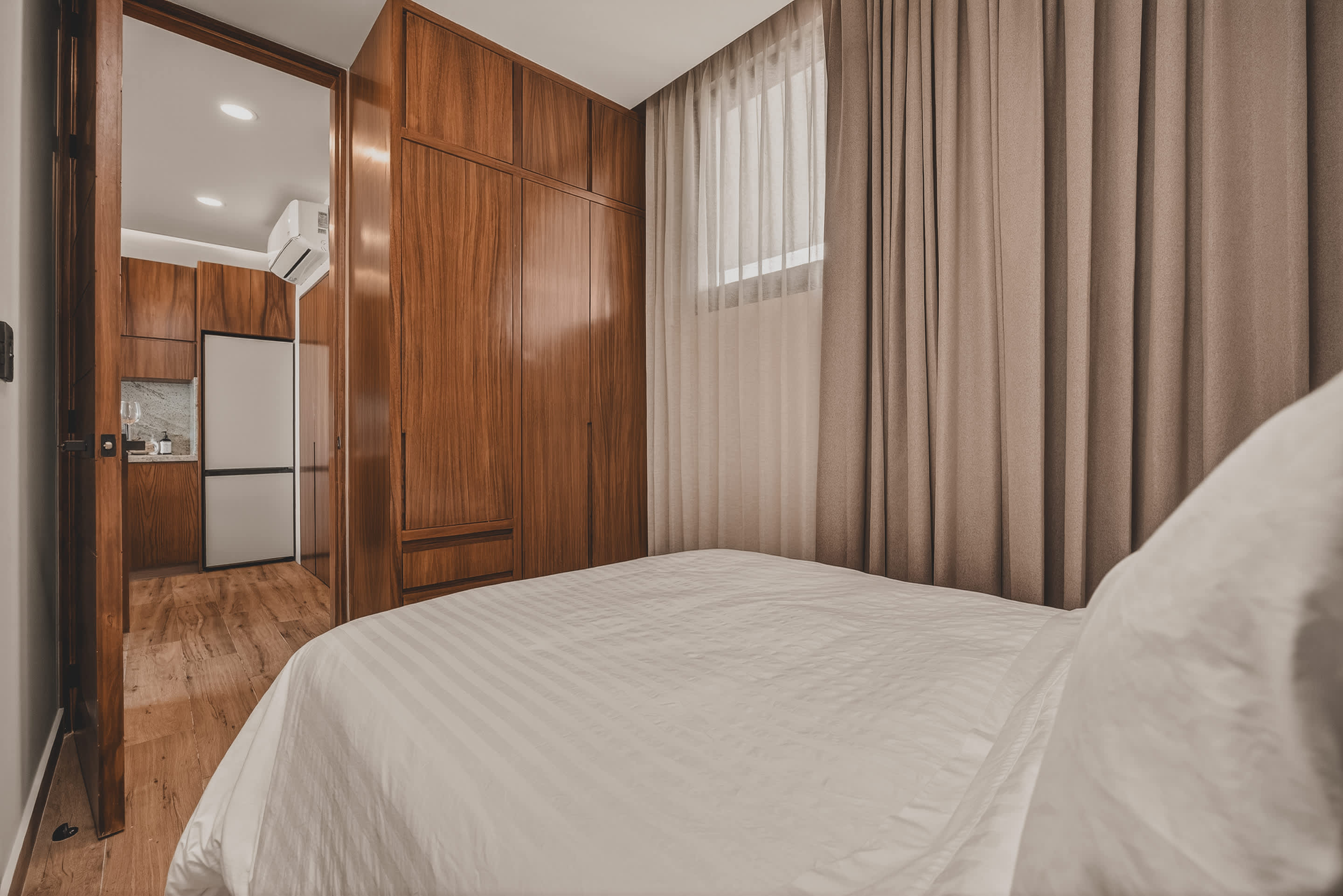 A cozy bedroom featuring a white-striped queen bed with a dark wooden headboard, complemented by warm wooden built-in wardrobes. A sheer and blackout curtain cover a window, and an open door reveals a modern kitchen area.