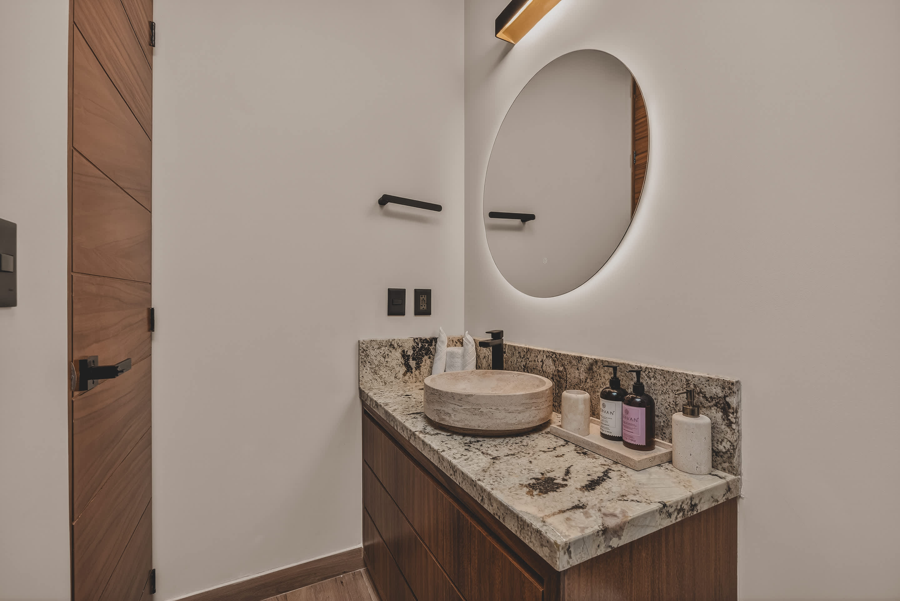 A close-up of the bathroom vanity highlights a stone vessel sink on a speckled granite countertop, flanked by modern black fixtures and various toiletries, beneath a backlit circular mirror.